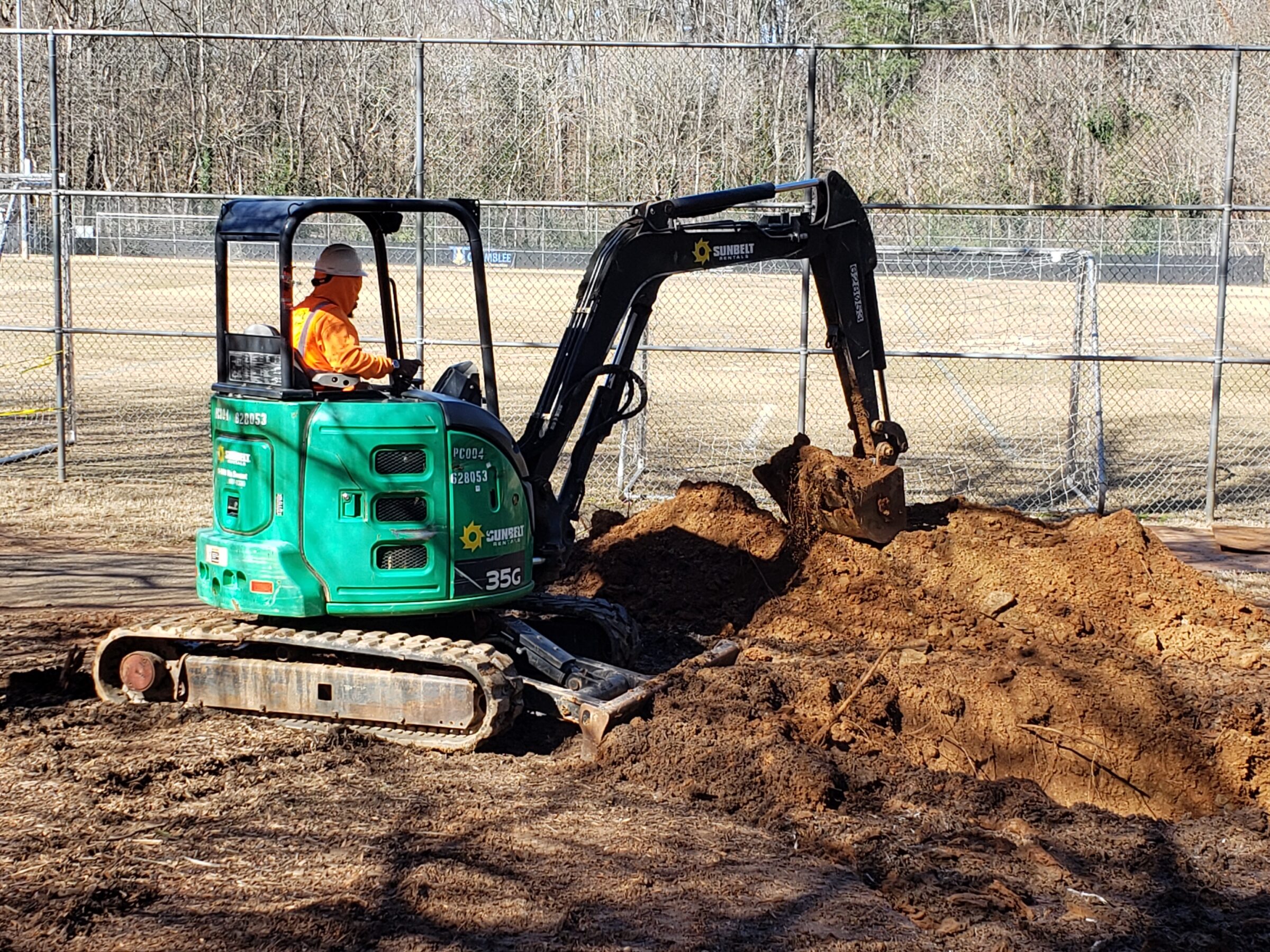 Una persona con chaleco naranja opera una excavadora verde cavando tierra, rodeada por una cerca de alambre y árboles al fondo.