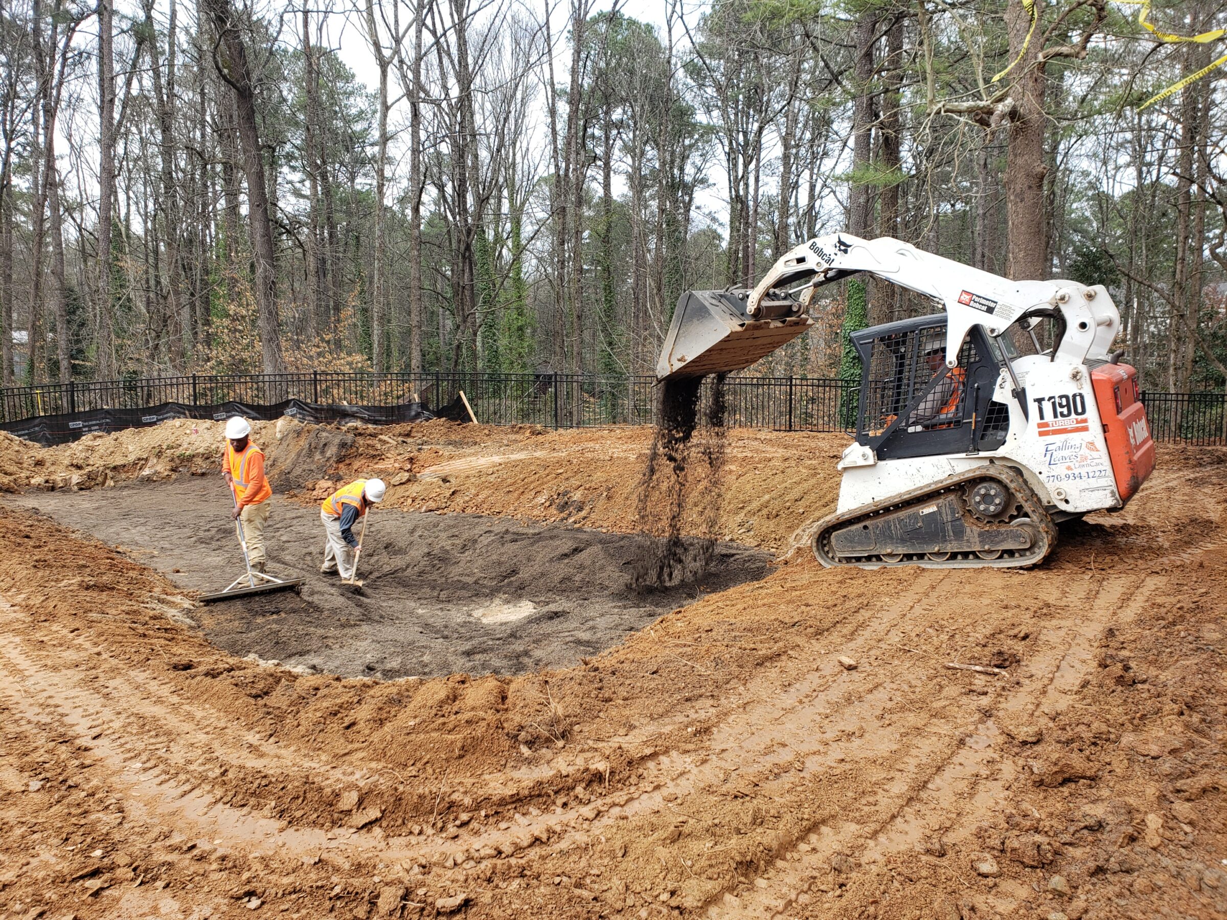 A construction site with two people in orange vests raking soil, and a skid steer loader depositing dirt; surrounded by trees.