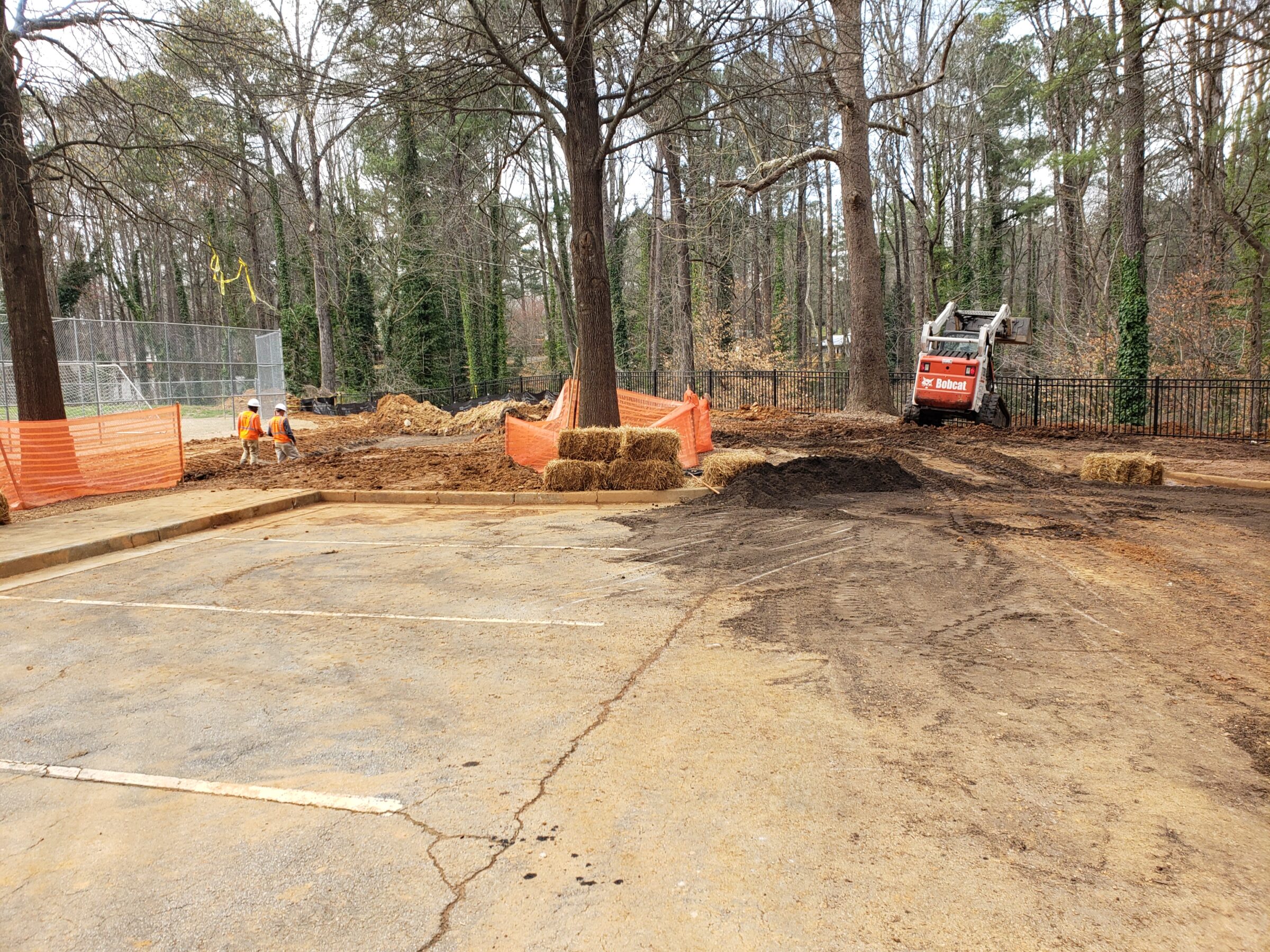 Two people in safety gear work on a construction site with machinery, surrounded by trees and orange barricades.