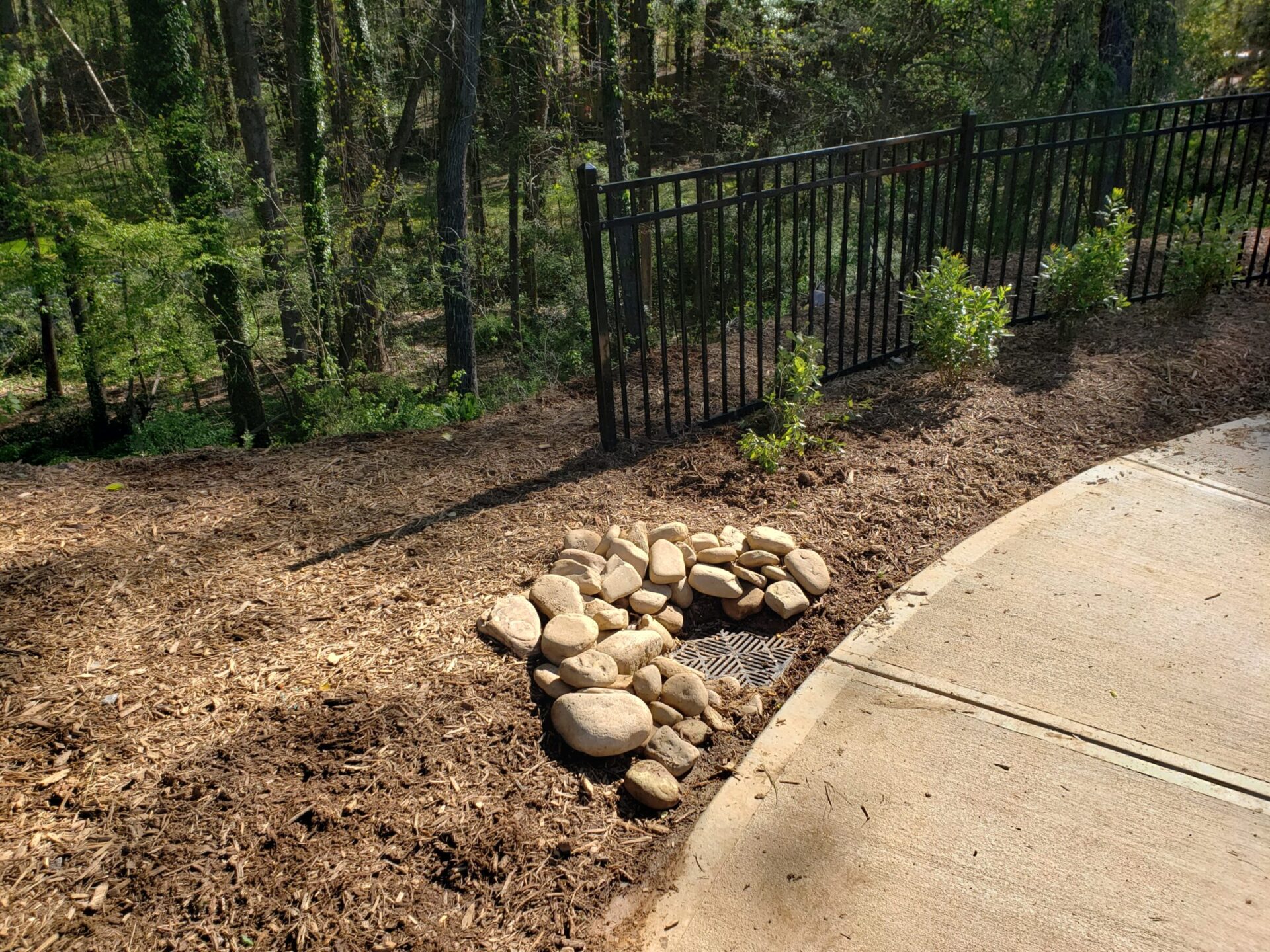 Sunny pathway with a black metal fence, adjacent mulch-covered area, and a stack of stones over a drainage grate near a forest.