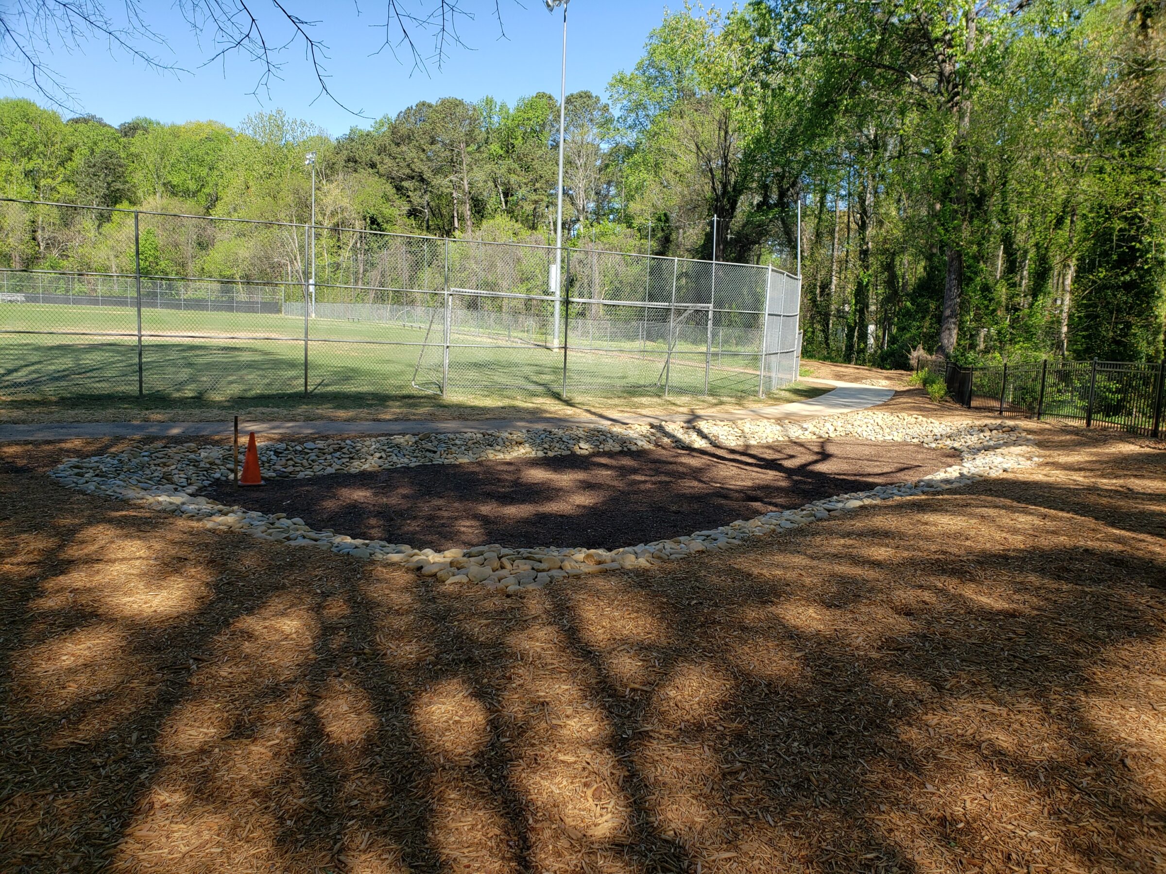 A sunny park scene shows a fenced baseball field, surrounded by trees. Pathway lined with stones; traffic cone stands in a gravel area.