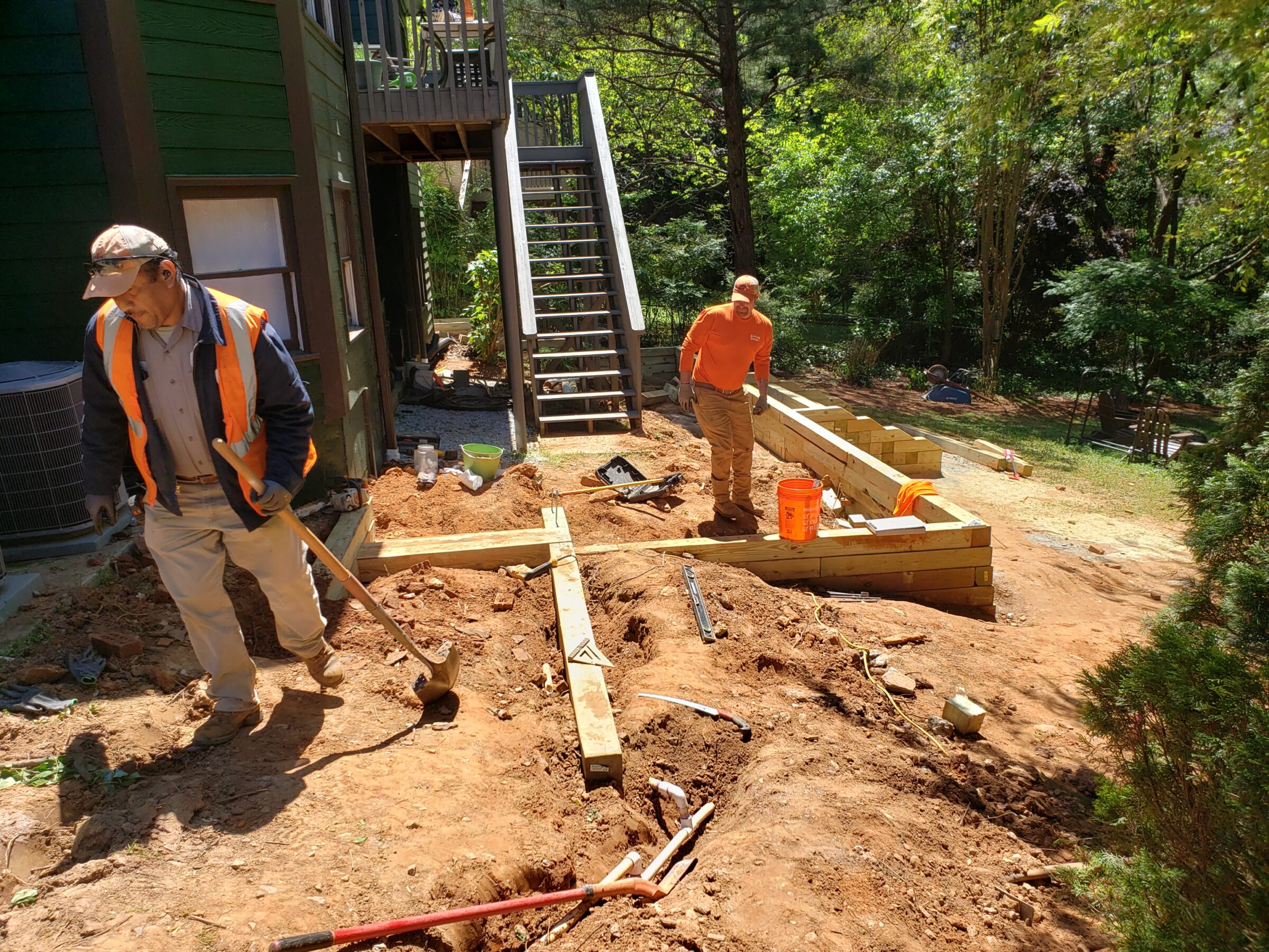 Two people work on construction near a wooden structure and stairs, surrounded by tools and greenery, under a clear sky.