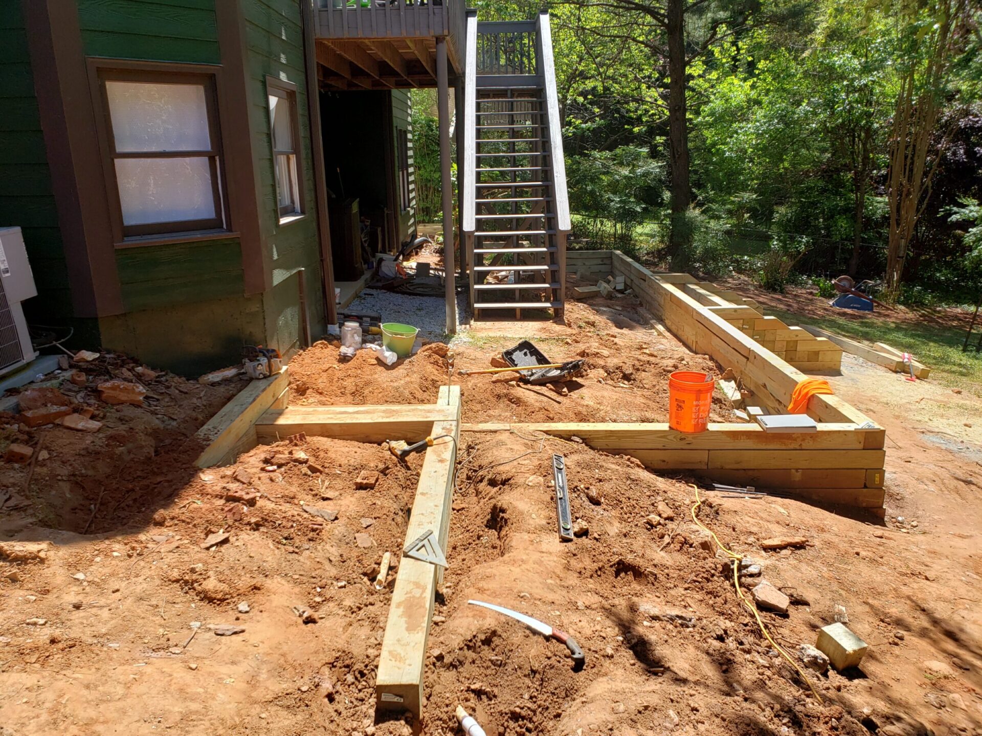 A backyard construction site with stairs, wooden frames, tools, and a bucket among partially dug soil, surrounded by greenery and trees.