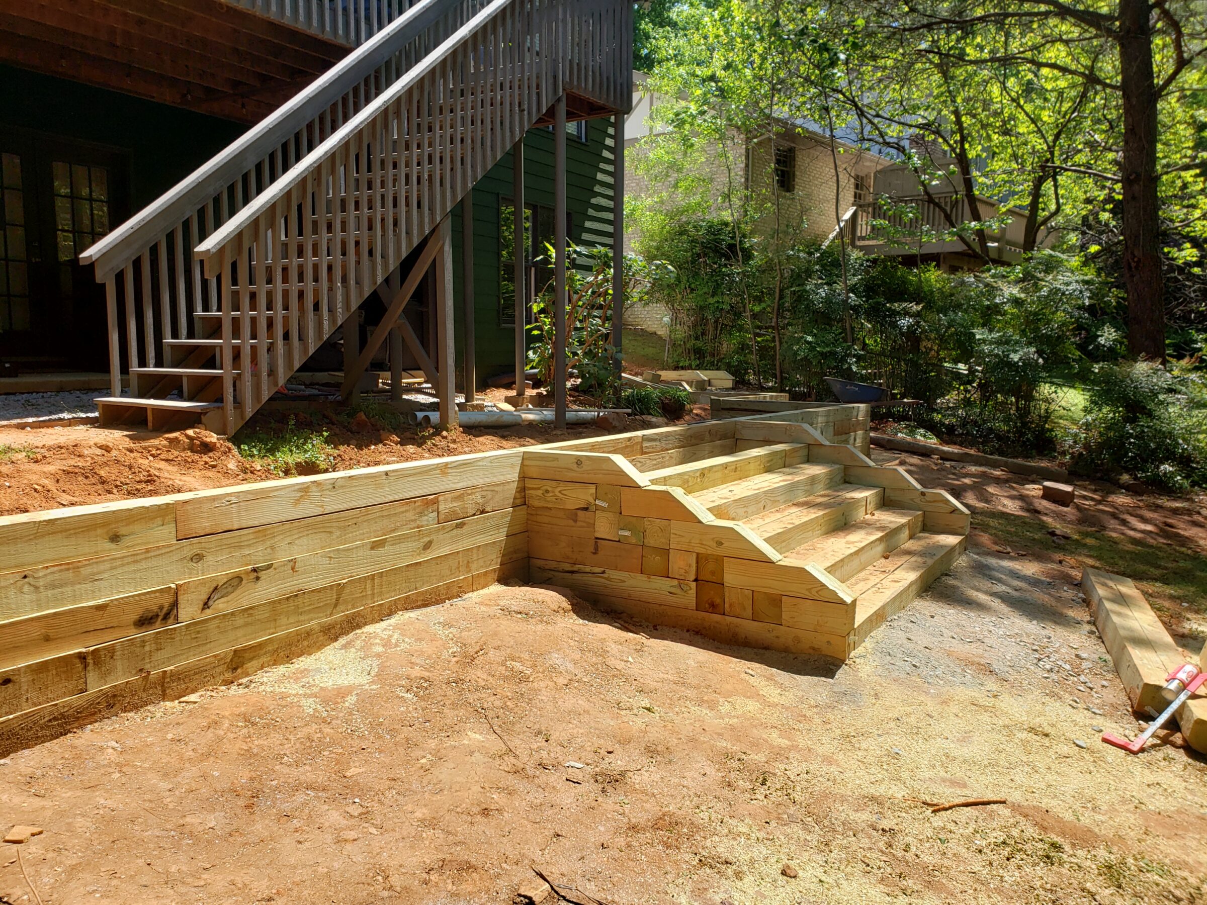 Wooden steps and retaining wall under construction, leading to elevated deck. Surrounding greenery and residential structures in background, under clear sky.