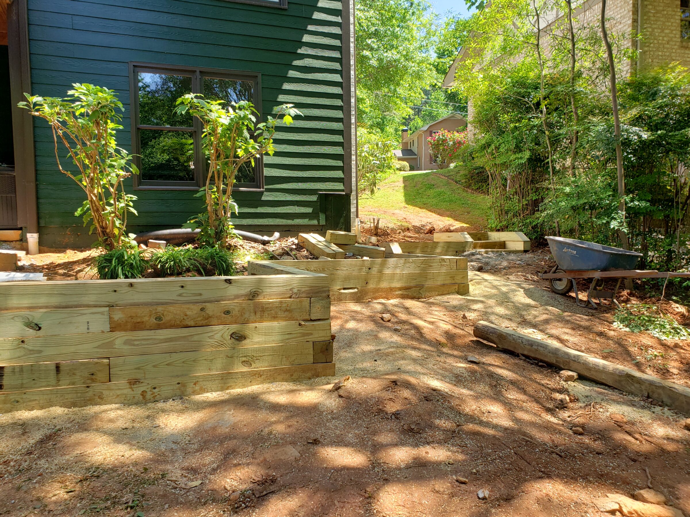 A green house with wooden retaining walls, surrounded by trees and shrubbery. A wheelbarrow rests nearby on the sunlit pathway.