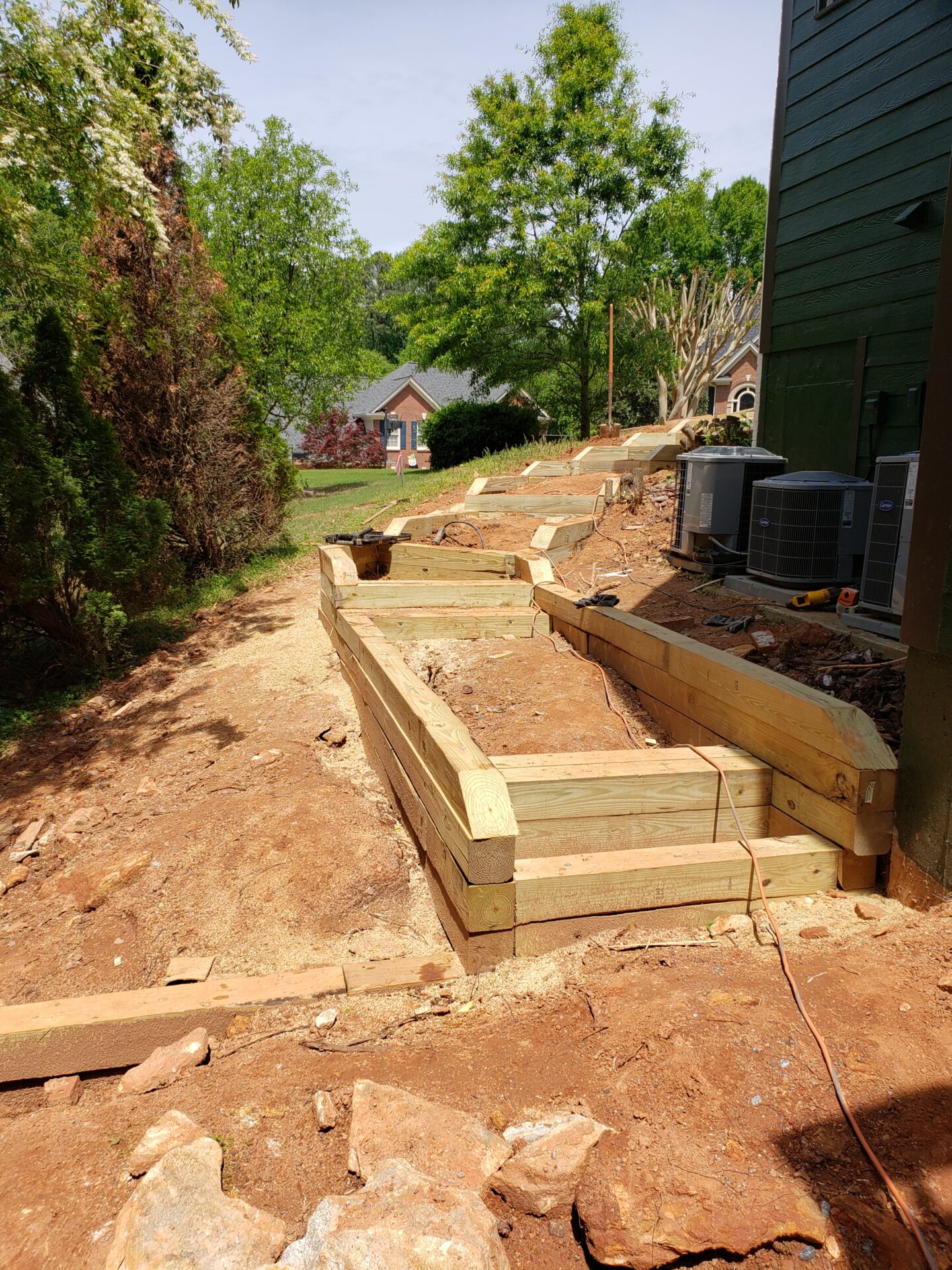 A backyard landscape features newly constructed wooden terraces, surrounded by trees and a neighboring house in the background under a clear sky.