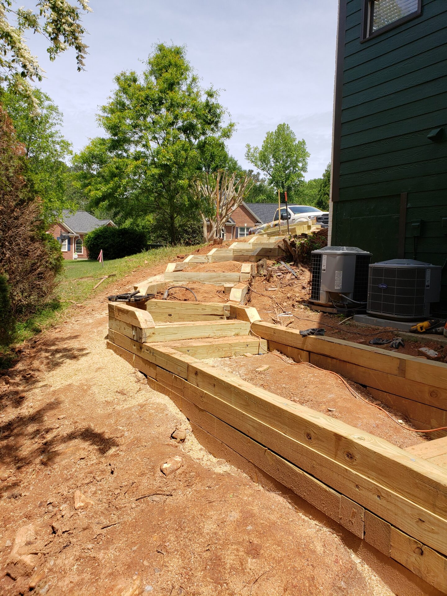 A wooden retaining wall under construction beside a house, surrounded by trees and neighboring homes in a residential area.