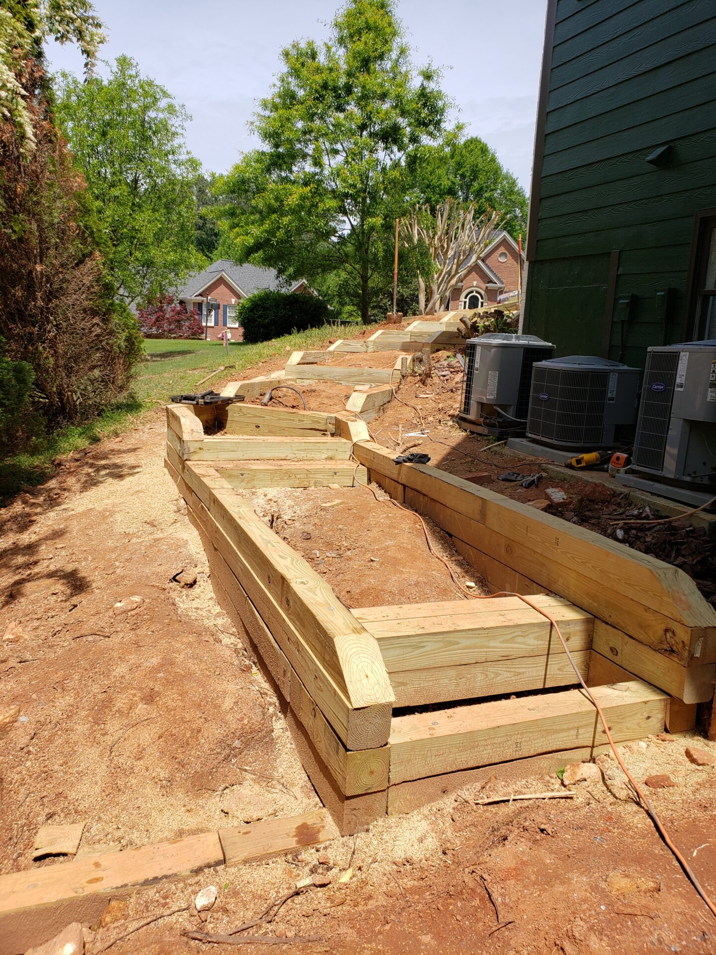 A newly constructed wooden retaining wall with steps, bordered by lush trees and a distant brick house, under a clear sky.