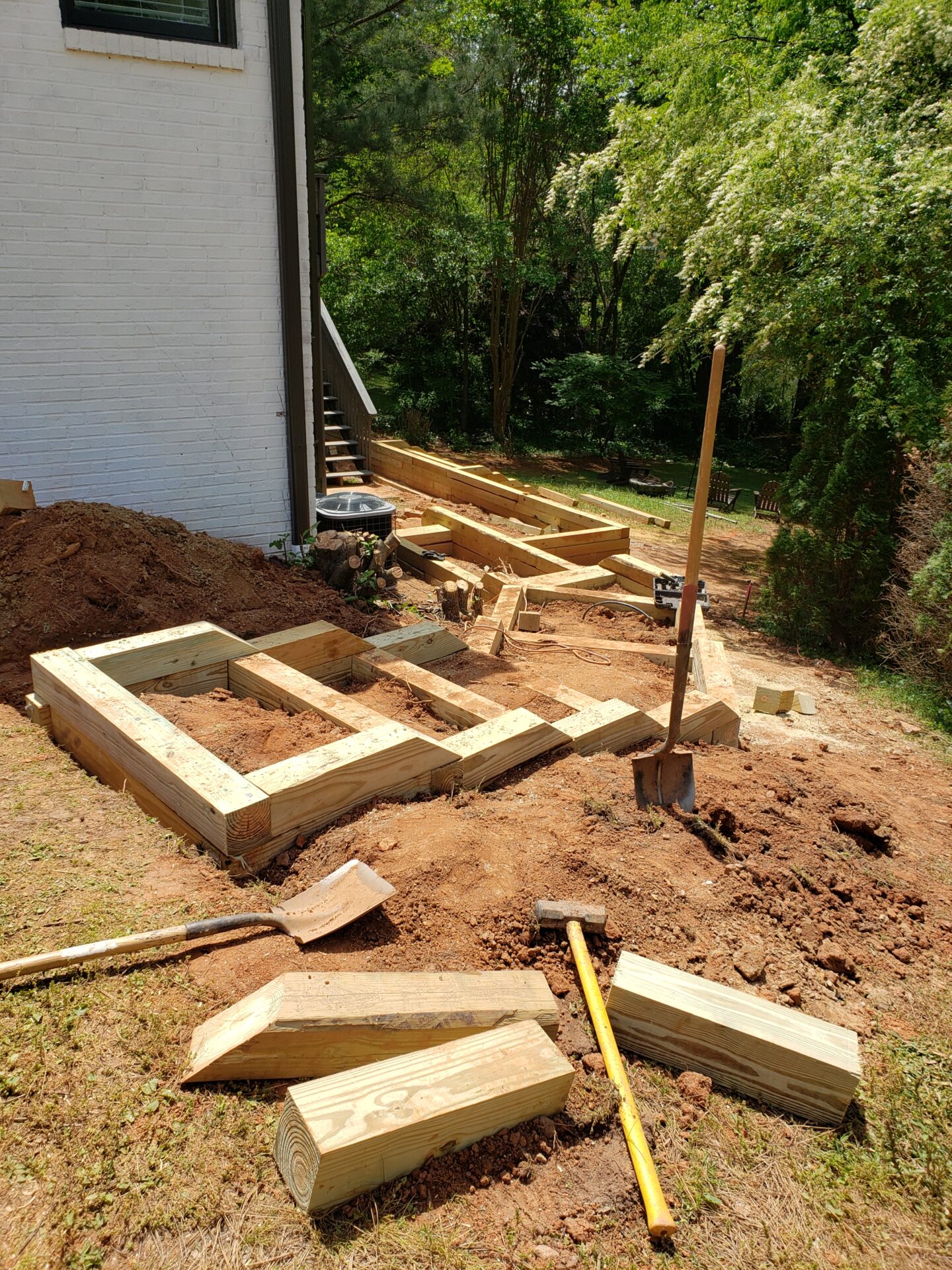 Construction site with lumber and tools beside a house, amidst a grassy area and trees, under bright sunlight. No people are present.