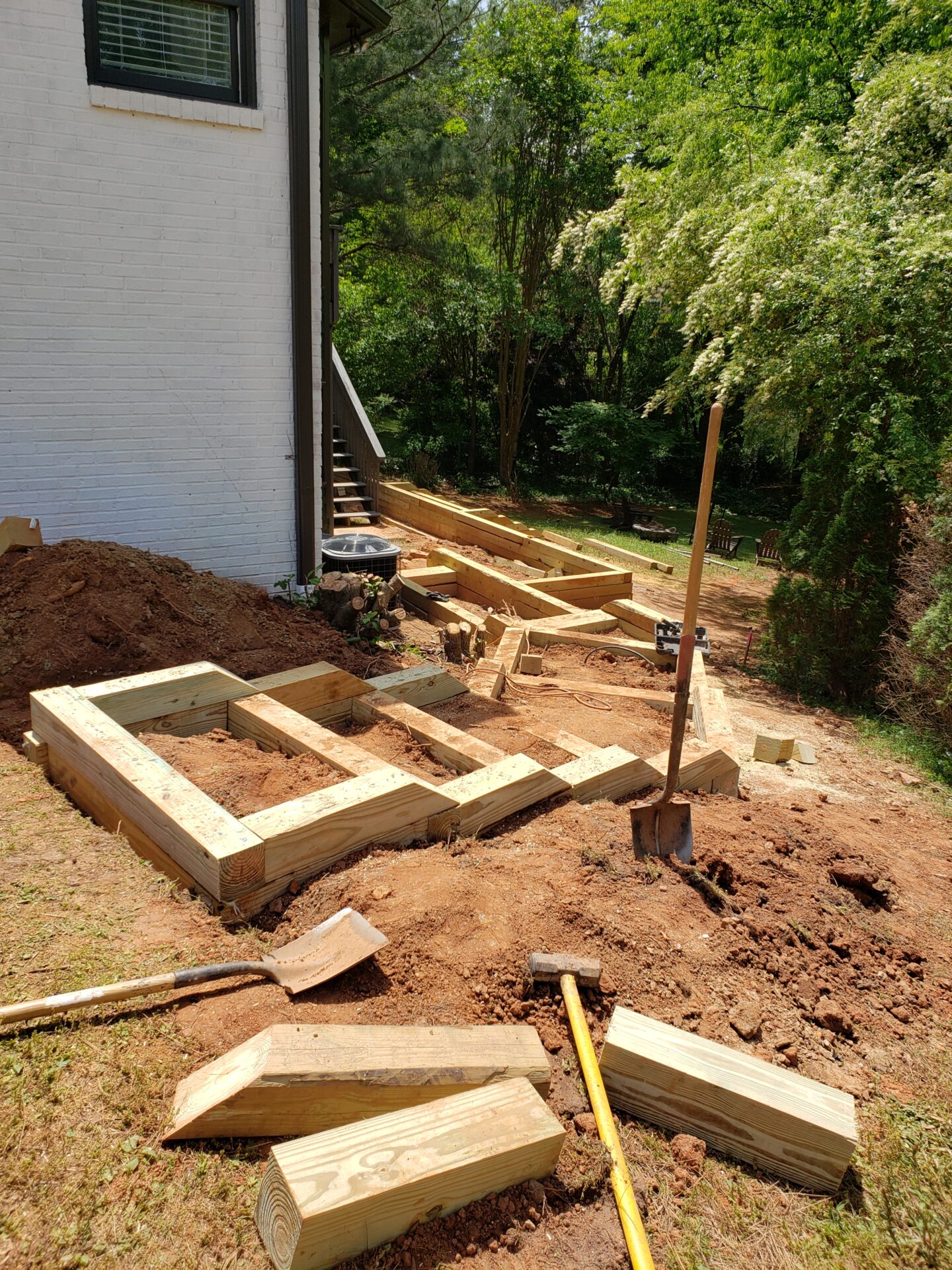 A construction site with wooden frames and tools near a white brick building, surrounded by trees and greenery on a sunny day.