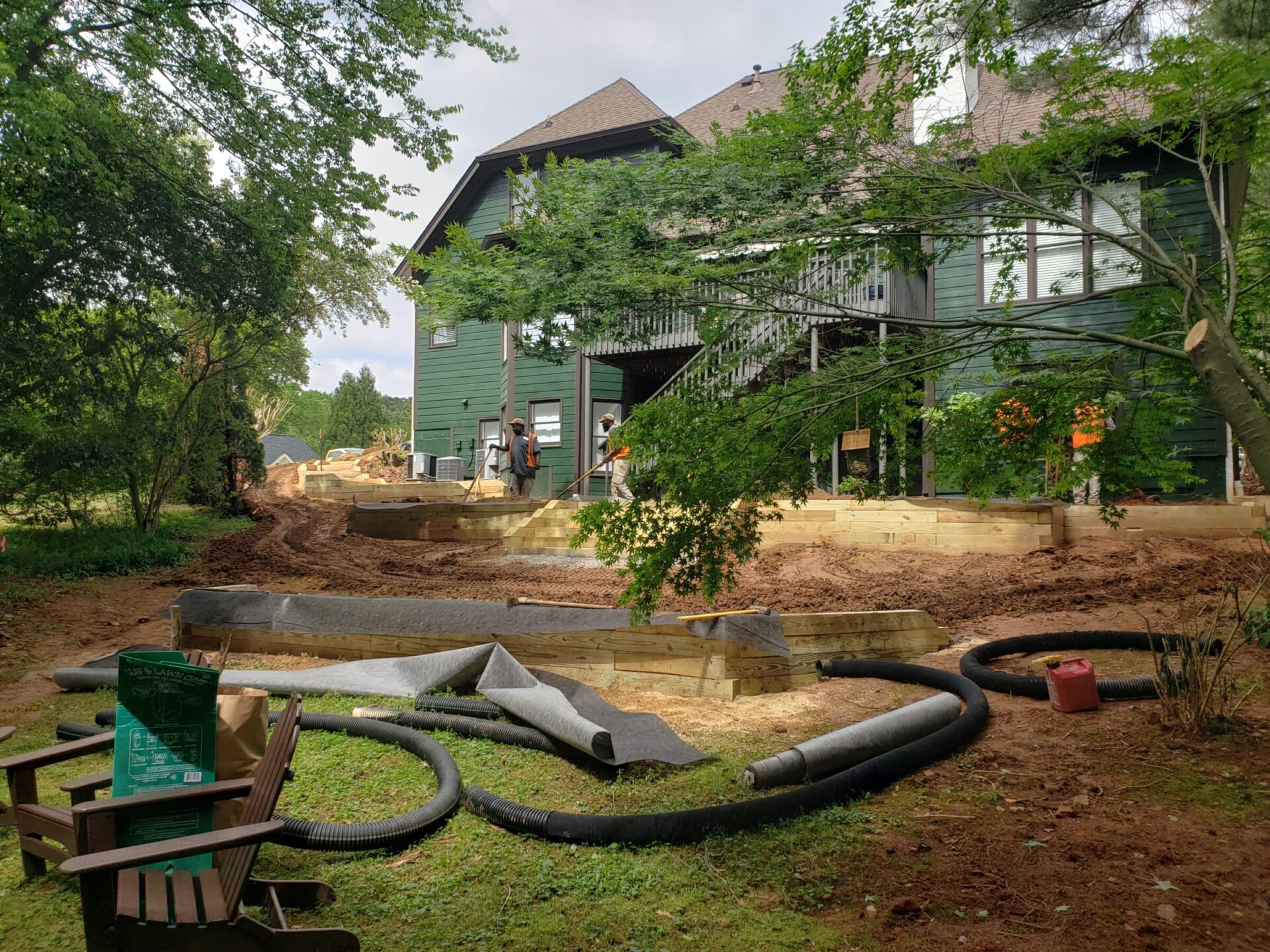 A house under construction with landscaping materials and two people working nearby, surrounded by trees and a lawn with chairs.