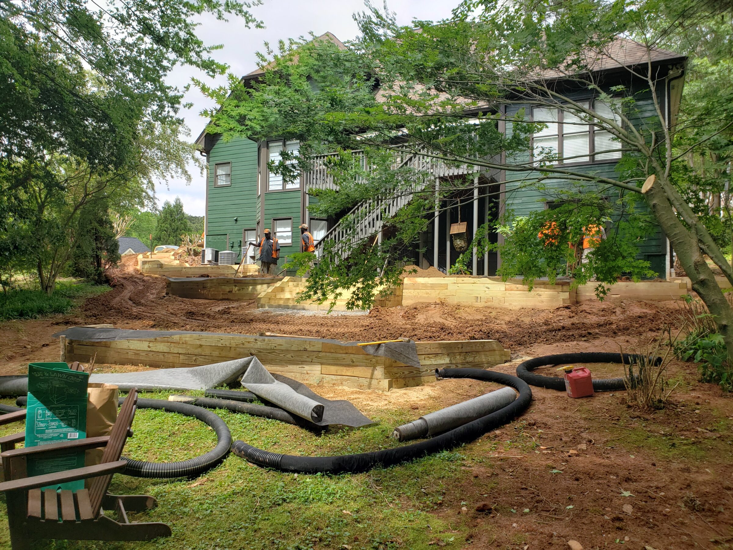 A green house under construction, with two people working outside. Wooden planks and construction materials are scattered around, surrounded by trees.