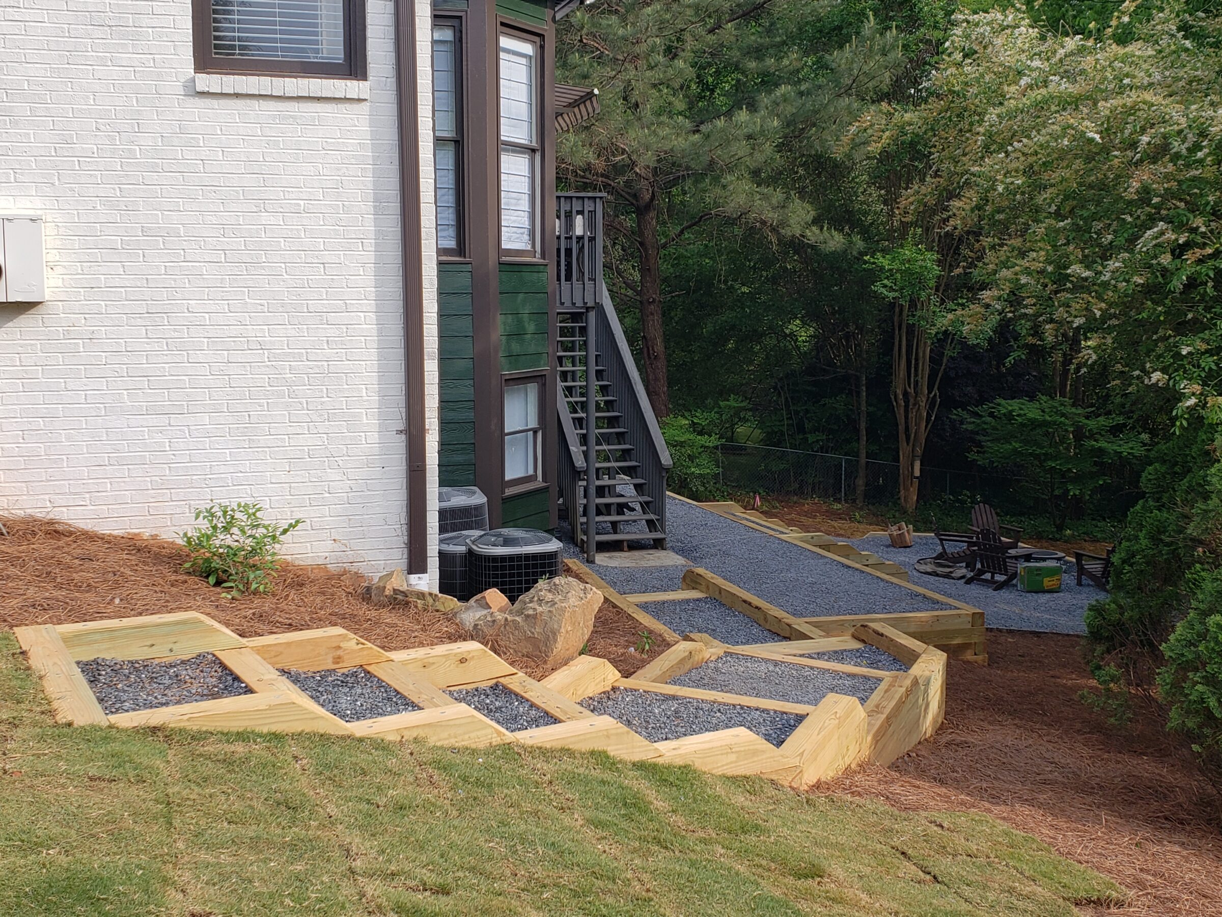 A house with white brick and green siding, gravel pathway, wooden steps, surrounded by trees and an outdoor seating area with fire pit.