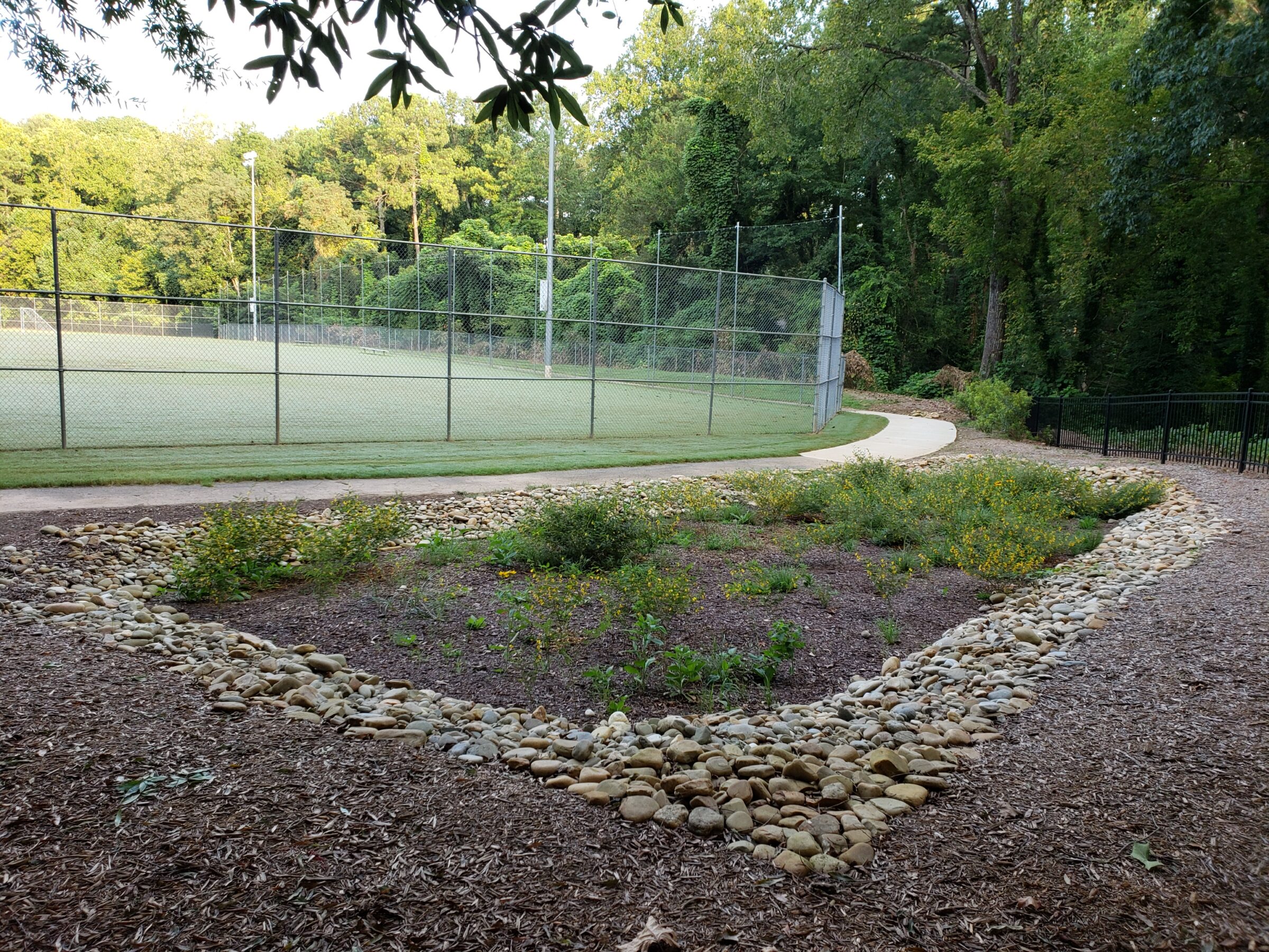 A baseball field fenced with chain-link, surrounded by lush trees and a garden bordered by stones, with a paved path.