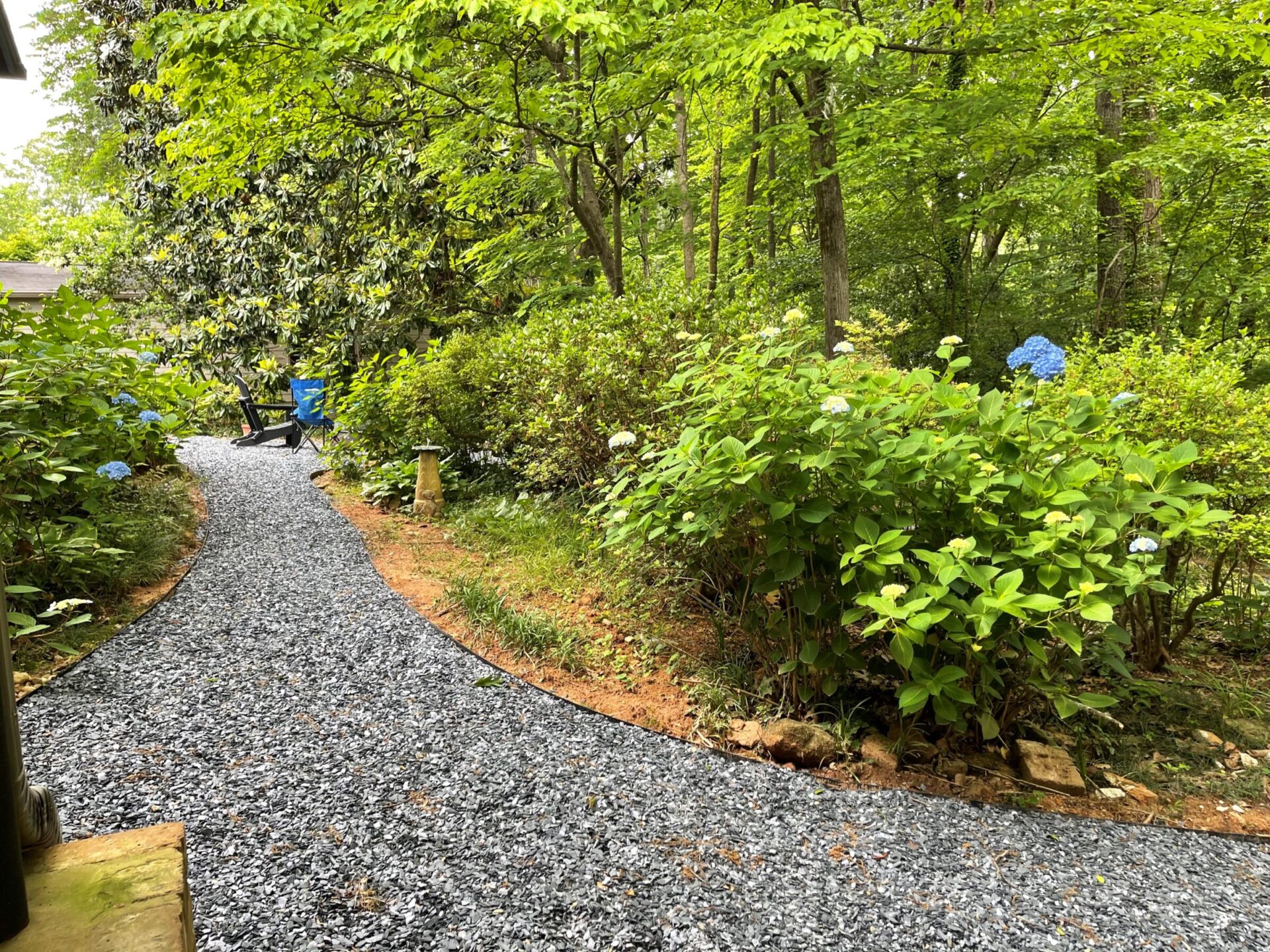 A gravel path winds through a green garden with hydrangea bushes and trees. A blue chair is visible beside the path.