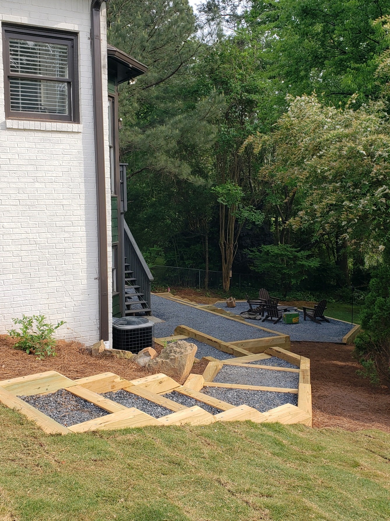 A backyard features a brick house, wooden steps, gravel pathways, and seating area amidst lush green trees and foliage.