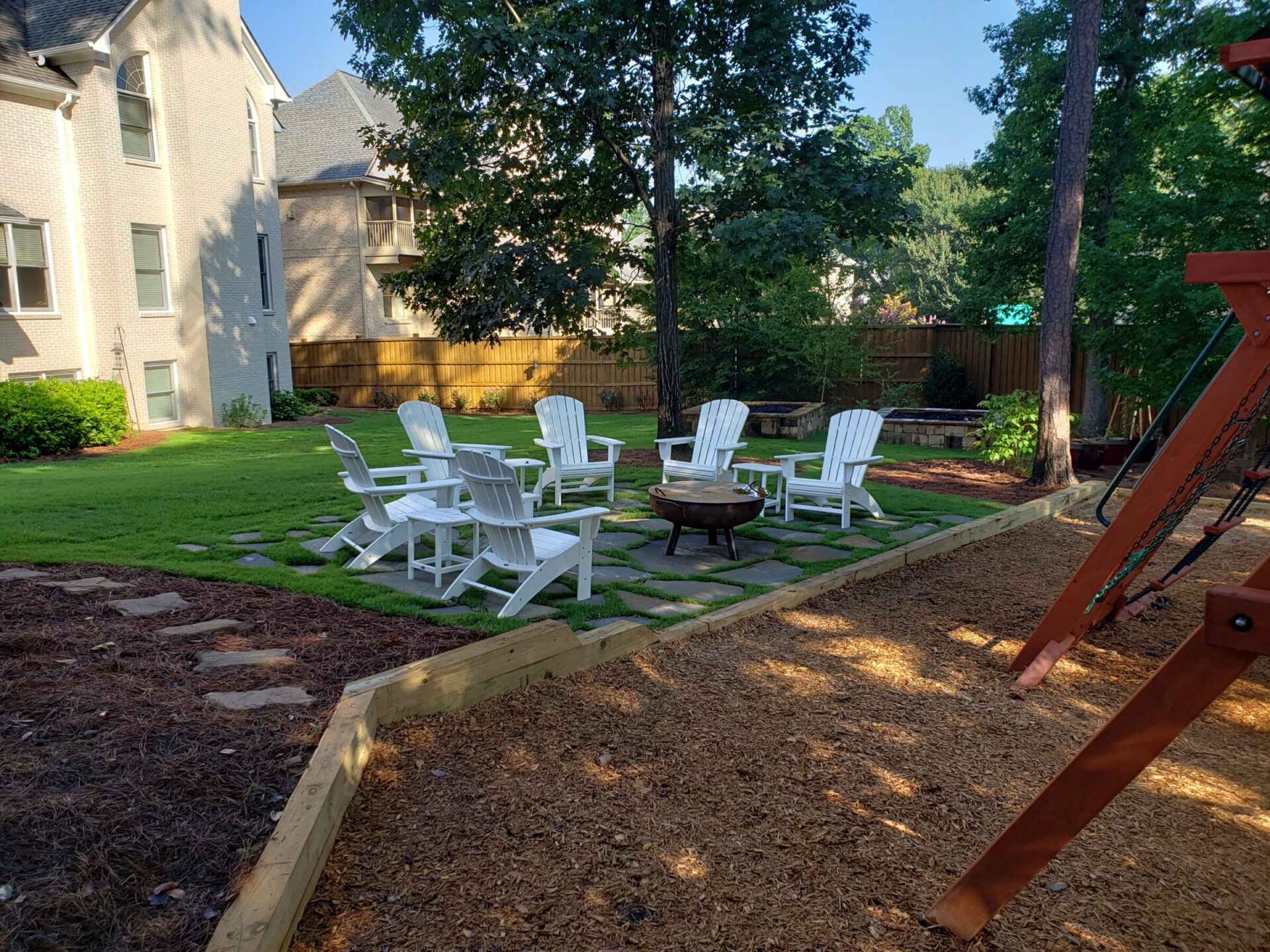 Backyard with white chairs around a fire pit, surrounded by trees and grass, adjacent to a playset and residential buildings.