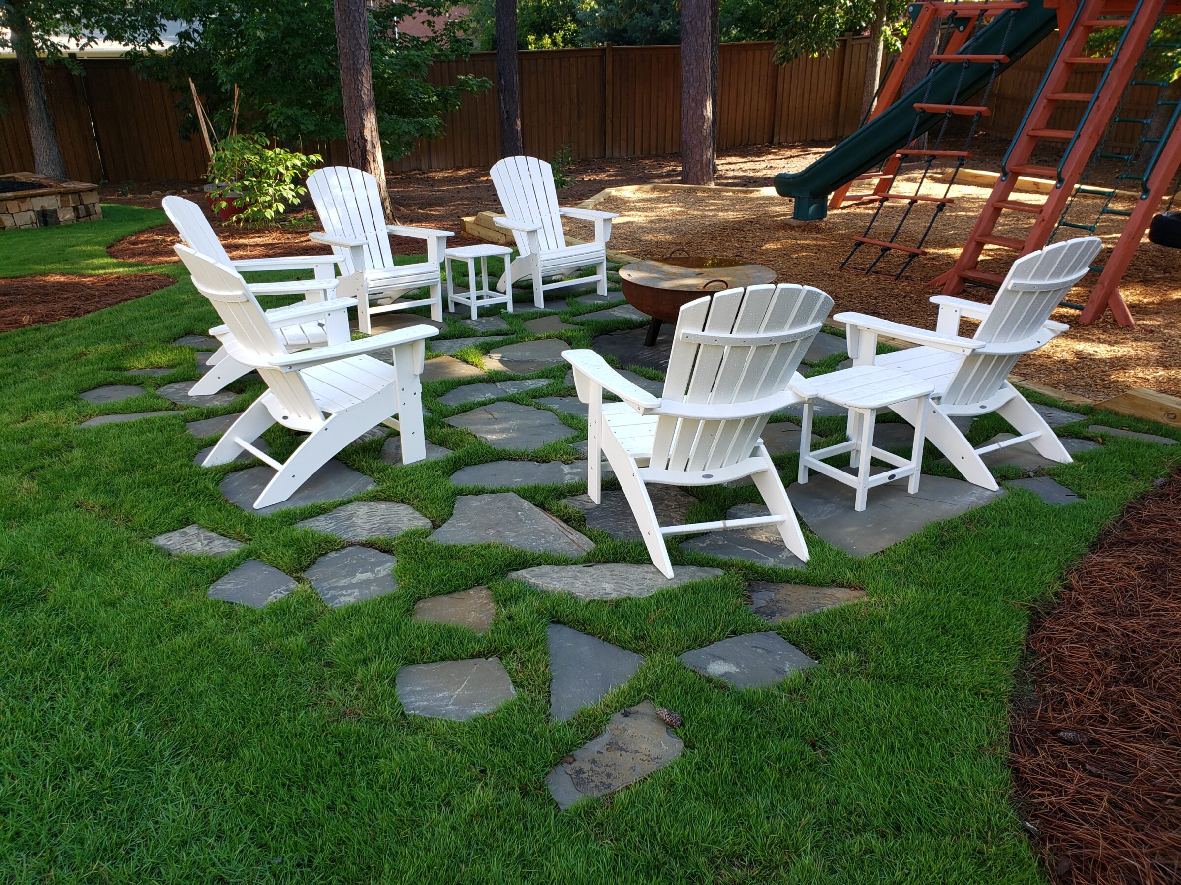 A backyard setting with white Adirondack chairs around a fire pit, surrounded by lush grass, stepping stones, and a wooden playset.