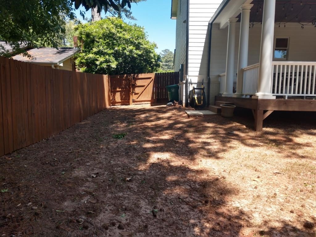 A shaded backyard with a wooden fence, porch, and trees. A gate and a few gardening tools are visible near the house.