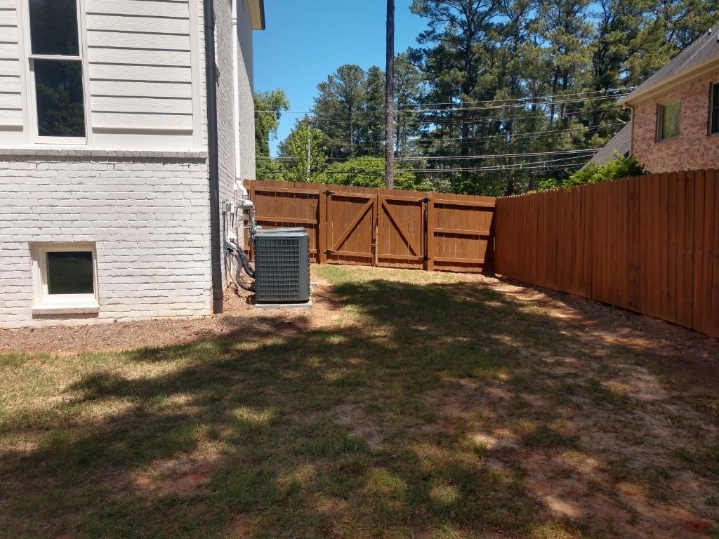 A well-maintained yard with grass, a white brick house, brown wooden fence, air conditioning unit, and tall trees in the background.
