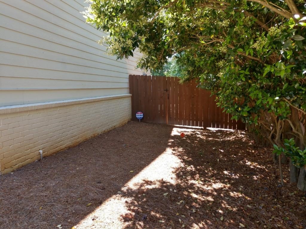A wooden fence and sign intersect with the corner of a brick house, shaded by leafy green trees, creating a secluded garden nook.