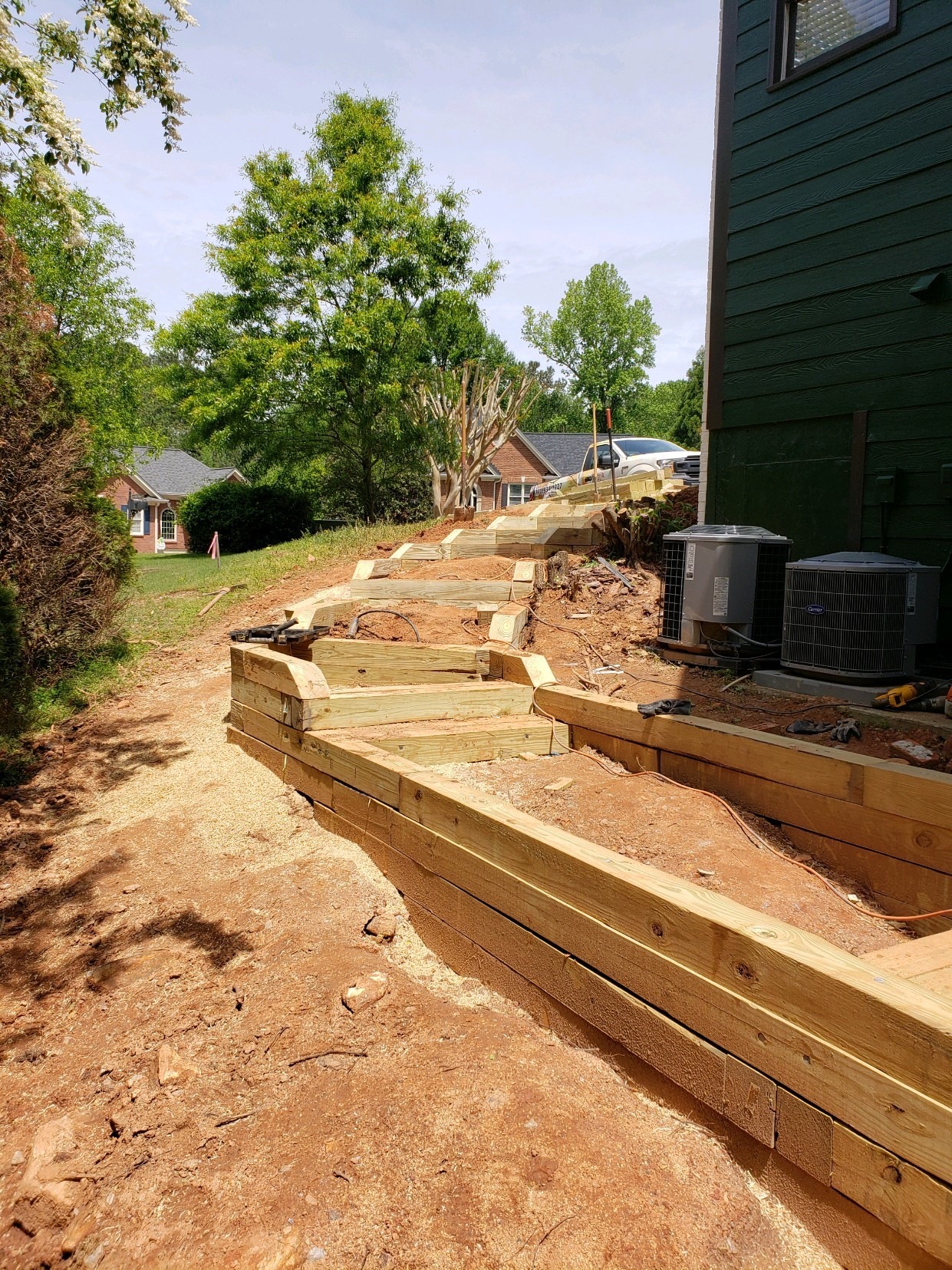 A sloped backyard shows new terraced wooden retaining walls under construction, with houses and trees visible in the background on a sunny day.