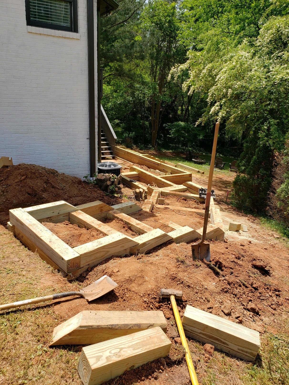 Wooden frame construction beside a white brick house, surrounded by green trees. Tools and lumber scattered on the dirt ground.