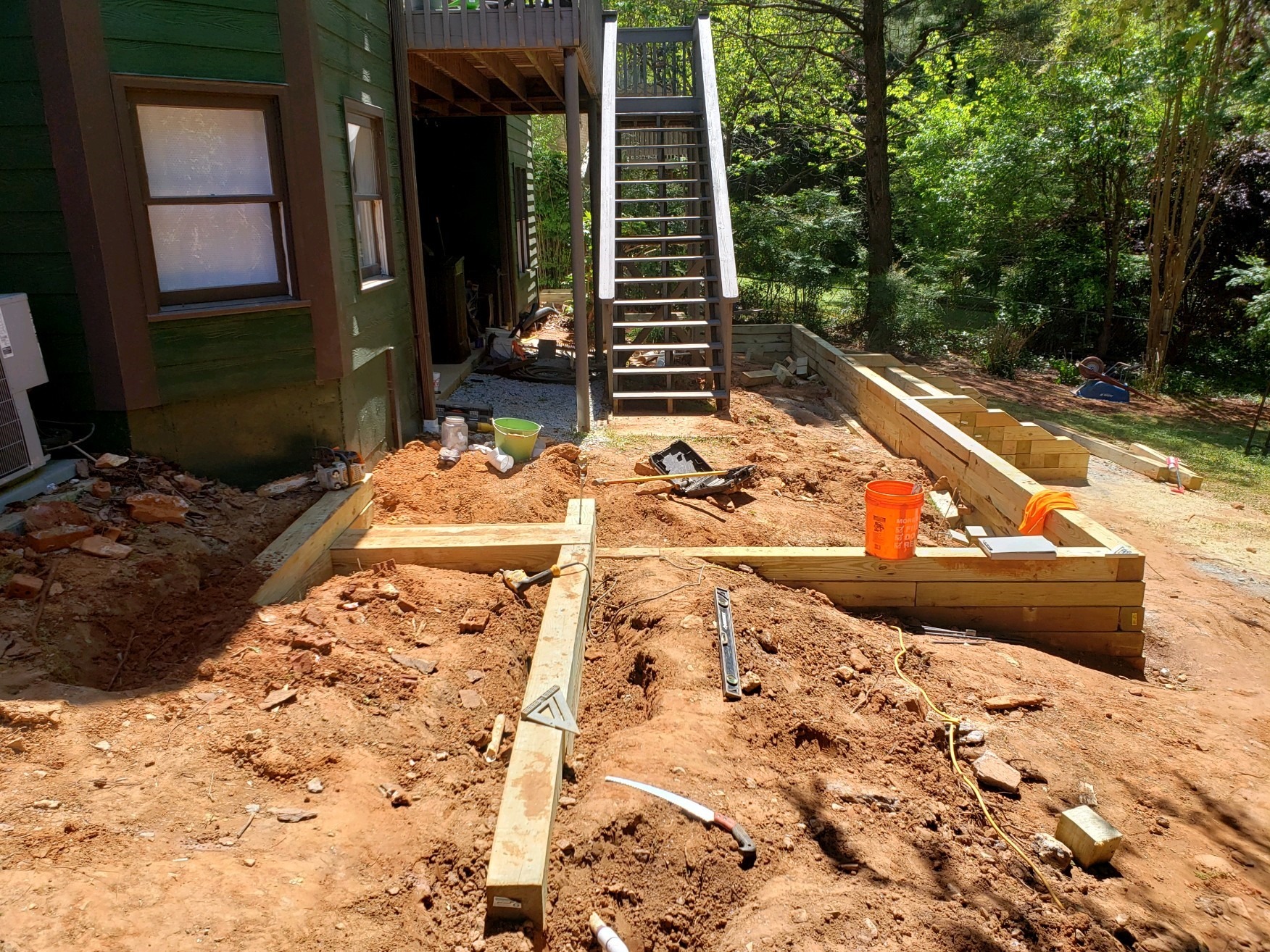 A construction site outside a green building features stairs, a wooden framework, and scattered tools, surrounded by dirt and trees in daylight.