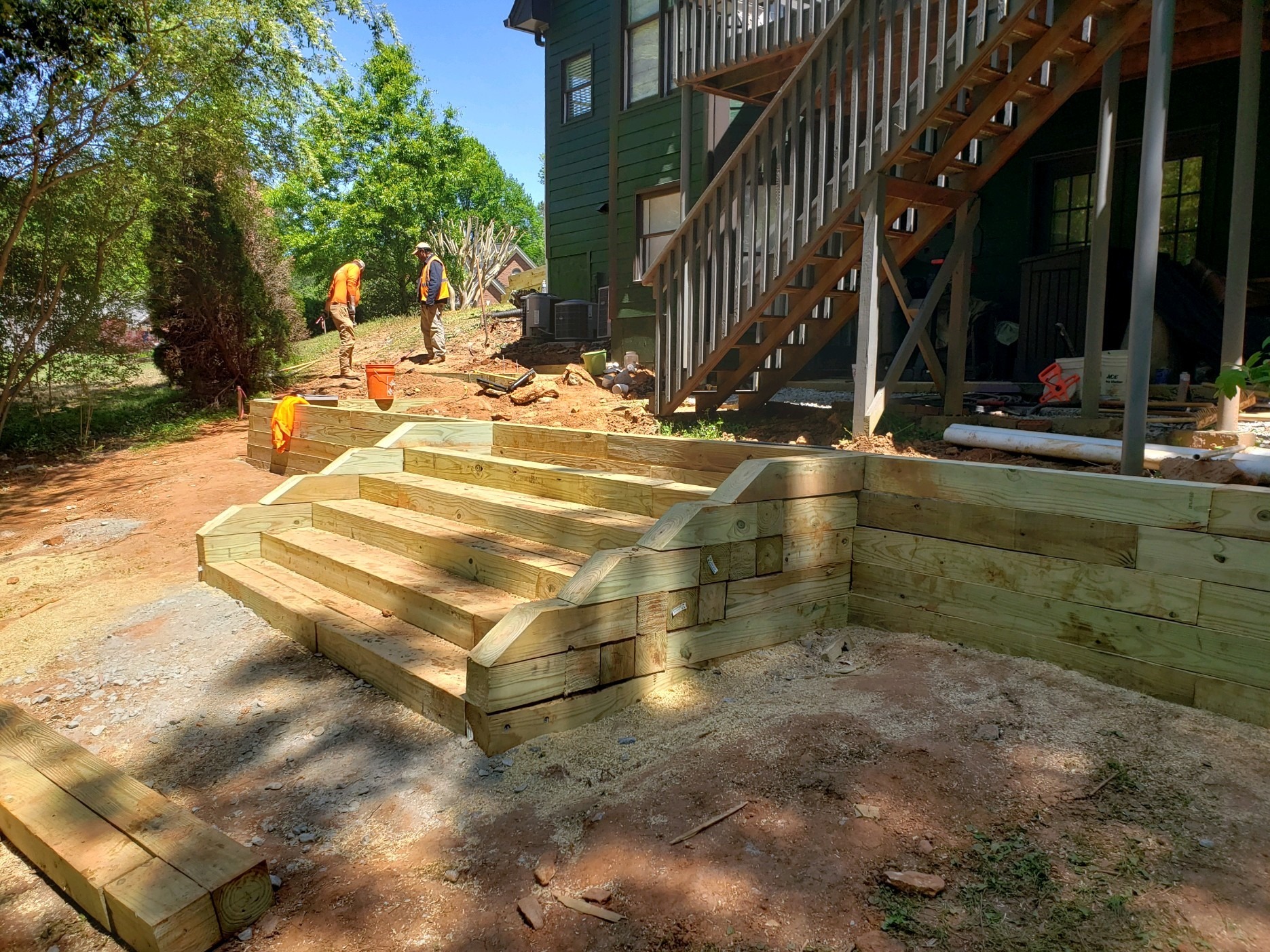 Two people in construction gear work on a wooden staircase and retaining wall outside a green building, surrounded by trees under a sunny sky.