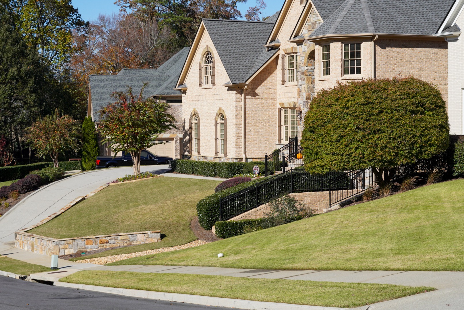 Suburban neighborhood with large brick house, manicured lawn, and neatly trimmed bushes. Driveway leads to attached garage. Person stands on walkway.