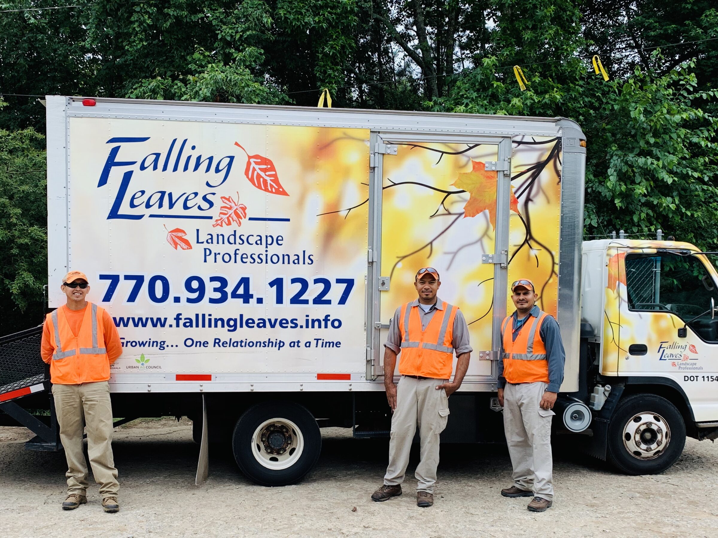 Three people in orange vests stand by a Falling Leaves landscaping truck with vibrant leaf design and contact details, surrounded by greenery.