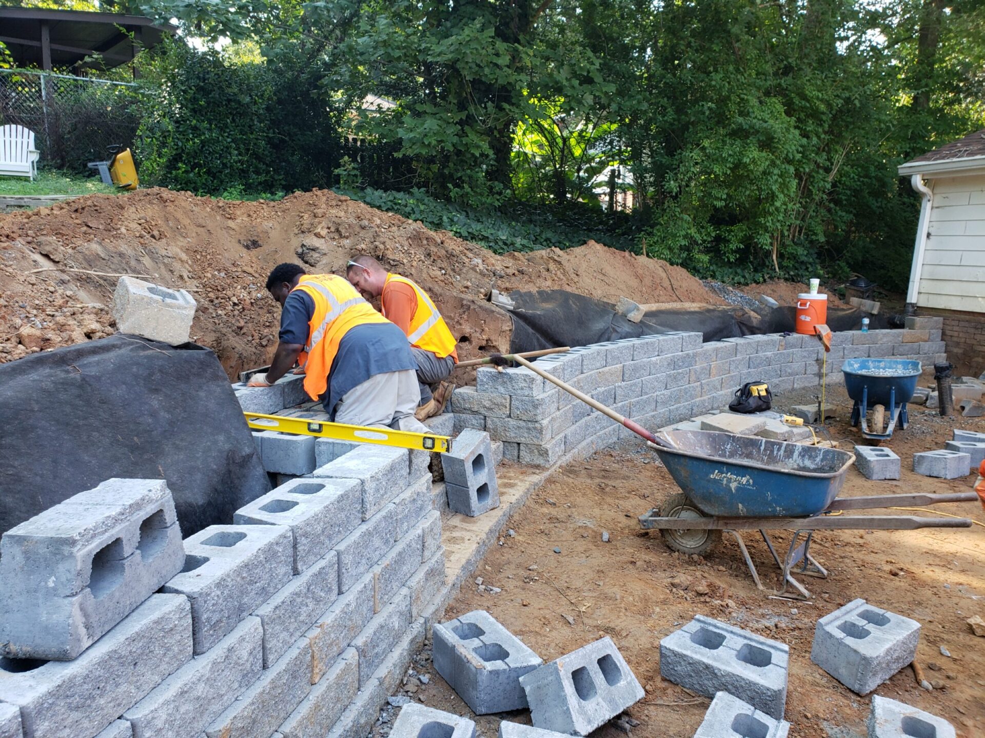 Dos personas con chalecos de construcción construyen un muro de contención de piedra en un patio trasero, rodeadas de herramientas y materiales, cerca de una casa blanca.