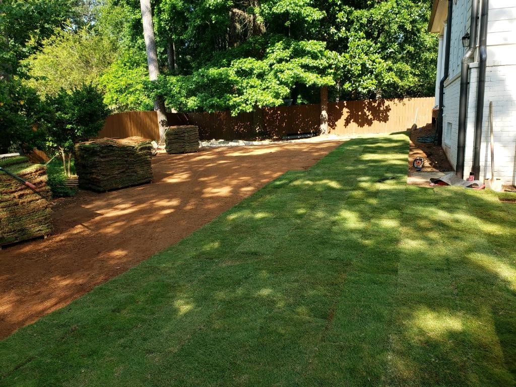 Rolls of sod are stacked beside a partially covered lawn area next to a house, surrounded by trees and a wooden fence.