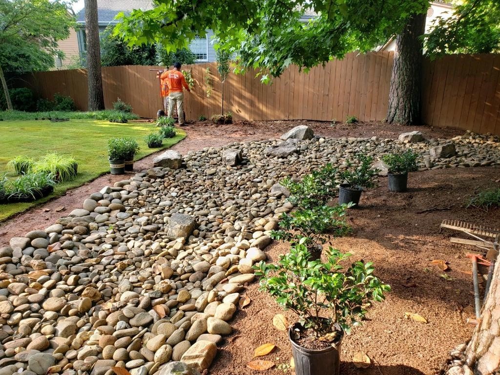Two persons work in a garden with rocky landscape, plants in pots, and a wooden fence. Trees provide shade in the background.