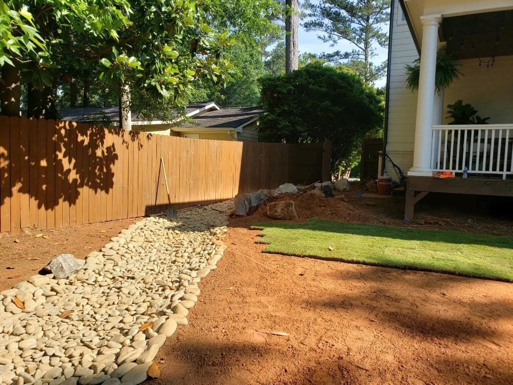A backyard features a wooden fence, dry riverbed with stones, grassy patch, porch with columns, and lush greenery under a clear sky.