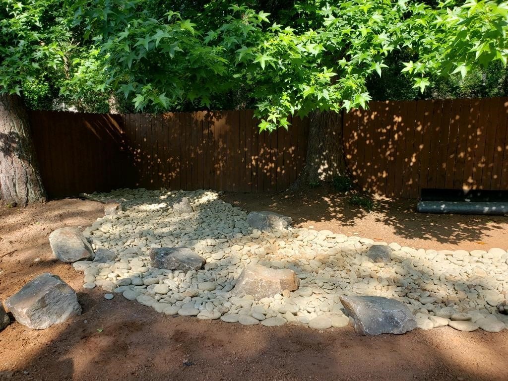 A serene garden with a dry riverbed of white stones and boulders, surrounded by lush trees, under a clear sky.