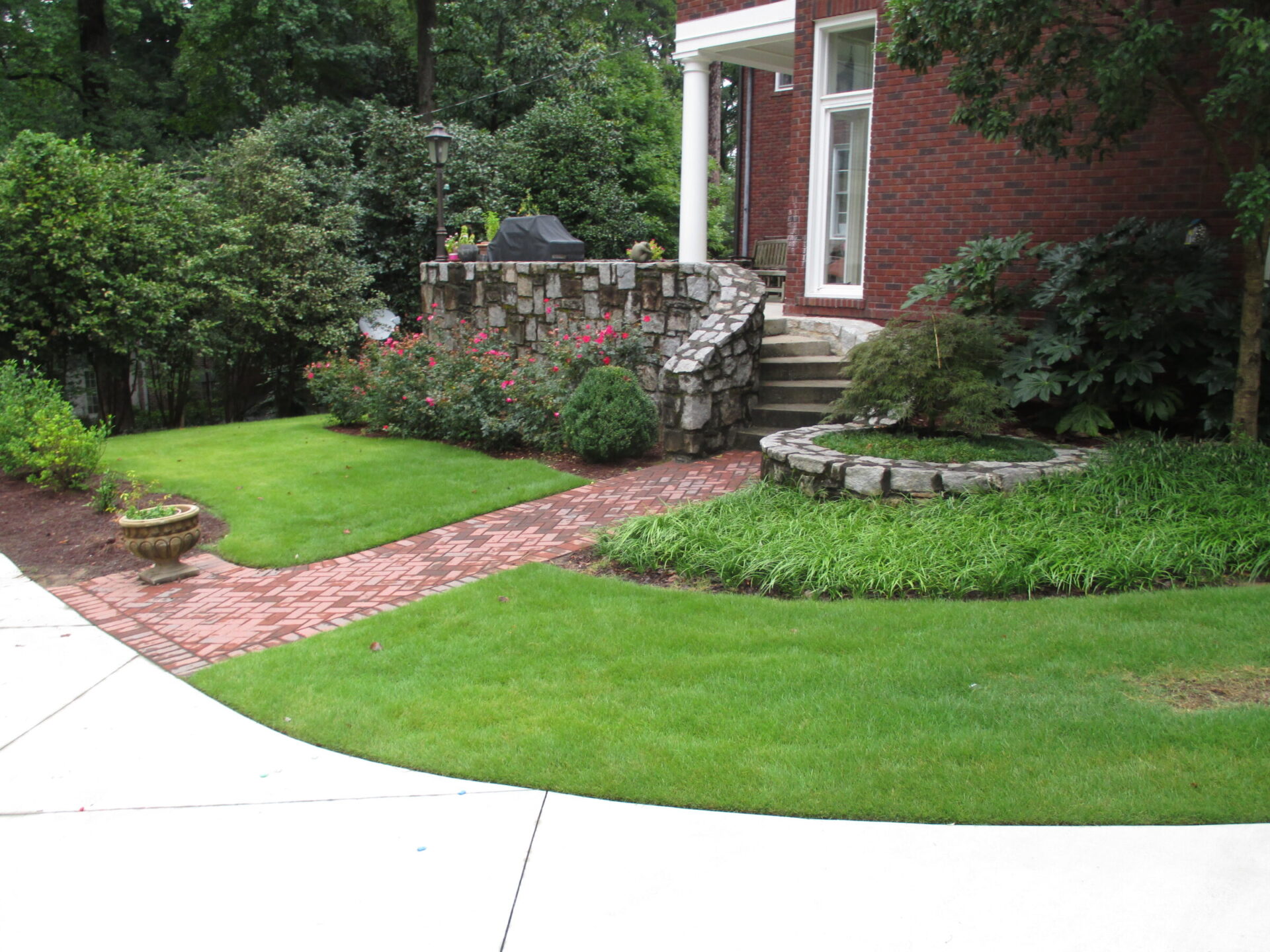 A landscaped garden with lush grass, brick pathways, stone walls, and shrubs surrounds a brick building with large windows.