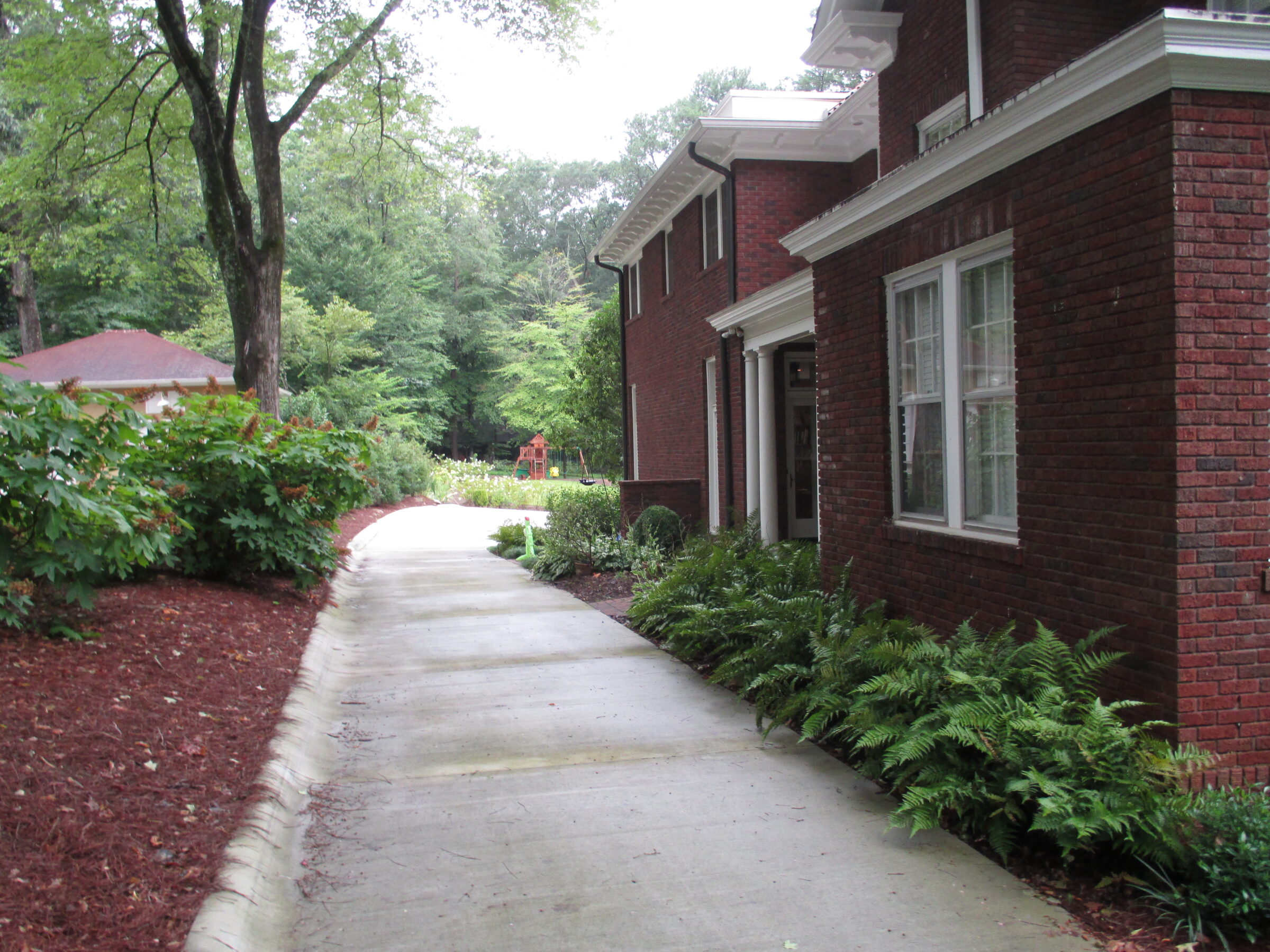 A curved concrete path leads past a red brick building, surrounded by lush greenery and tall trees, towards a red-roofed structure.