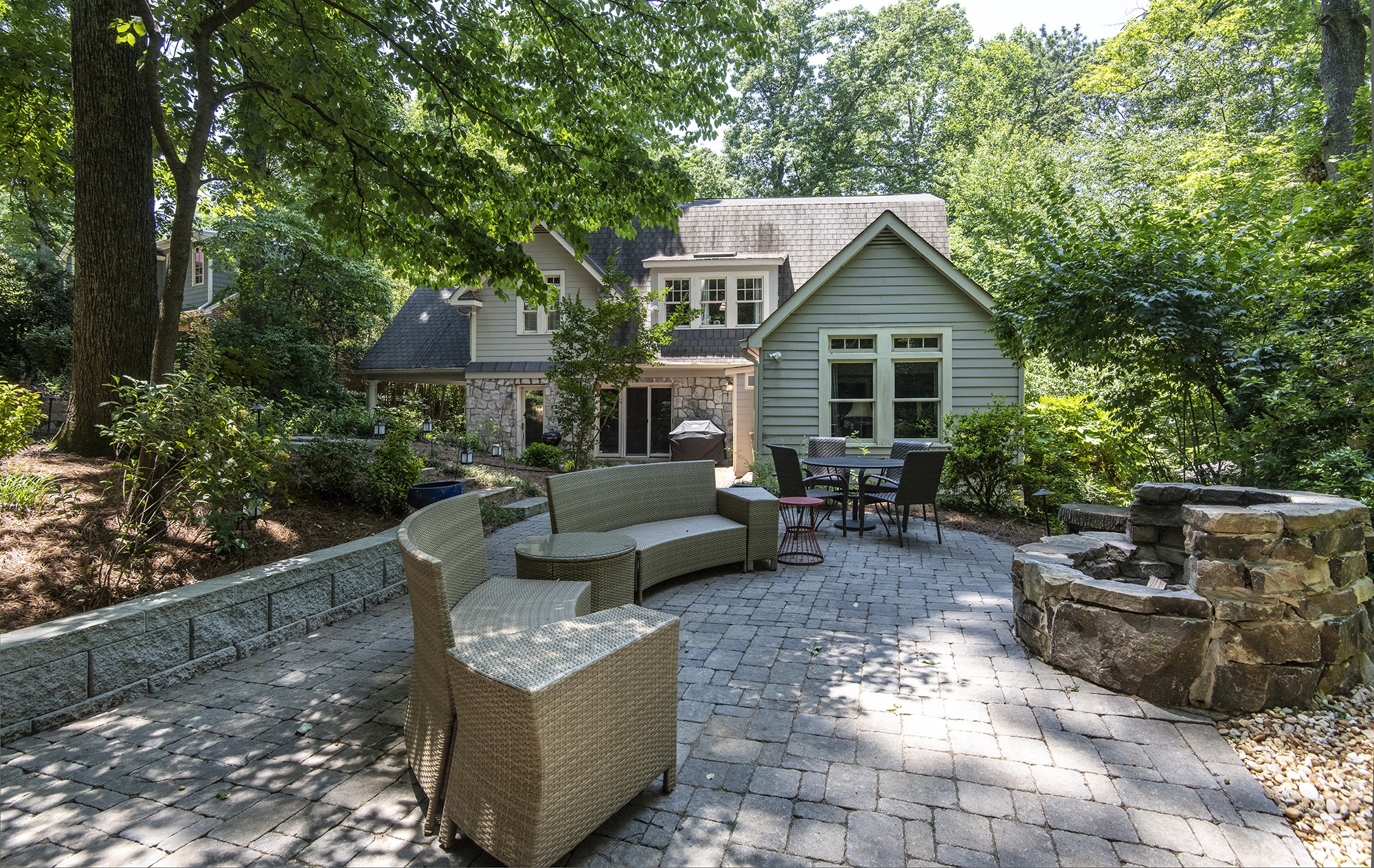 A serene backyard with patio furniture, stone fire pit, and a charming house surrounded by lush greenery and tall trees.