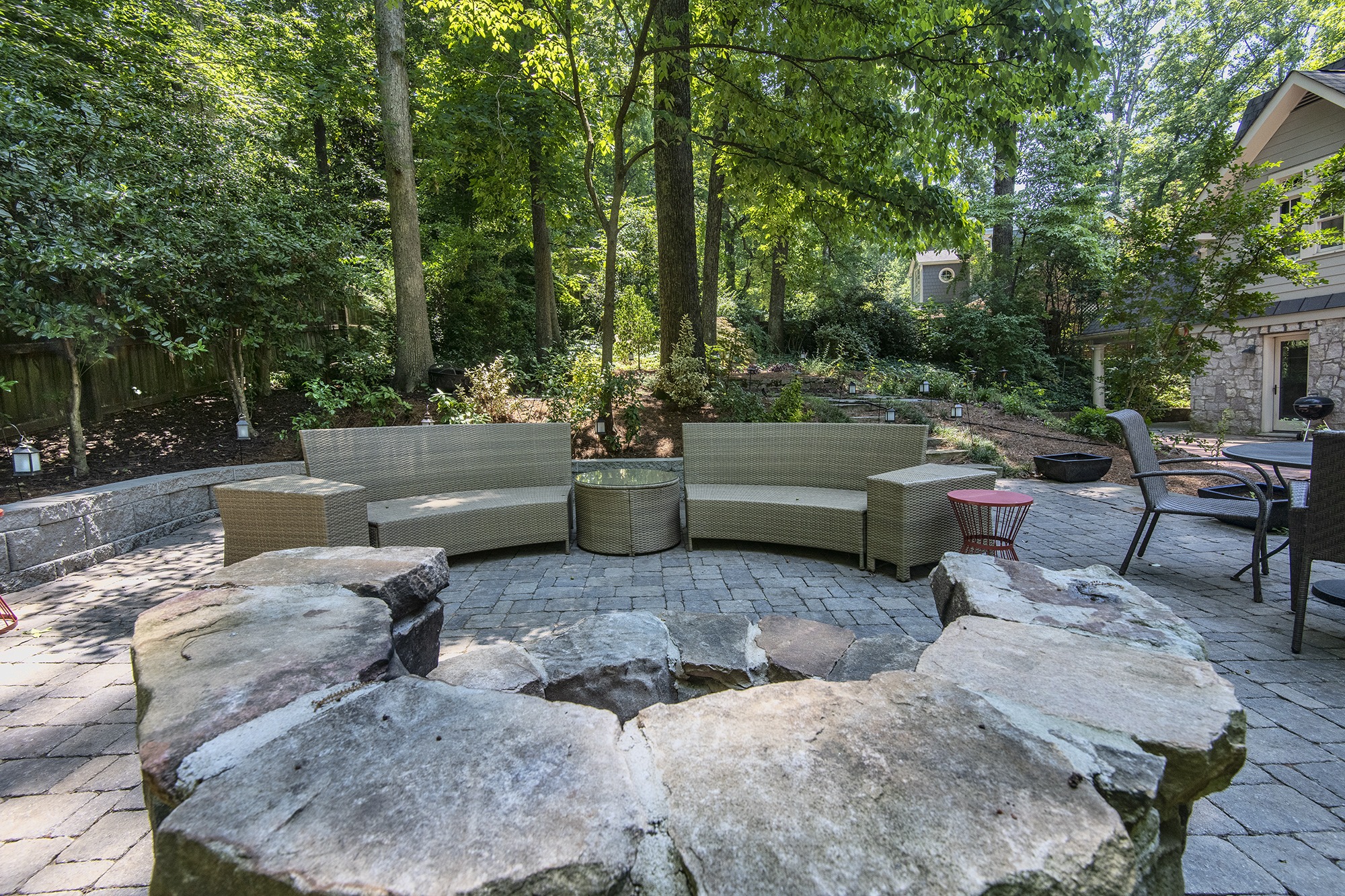 A cozy outdoor seating area with wicker furniture is set amidst lush greenery, surrounded by trees and a stone path, near a house.
