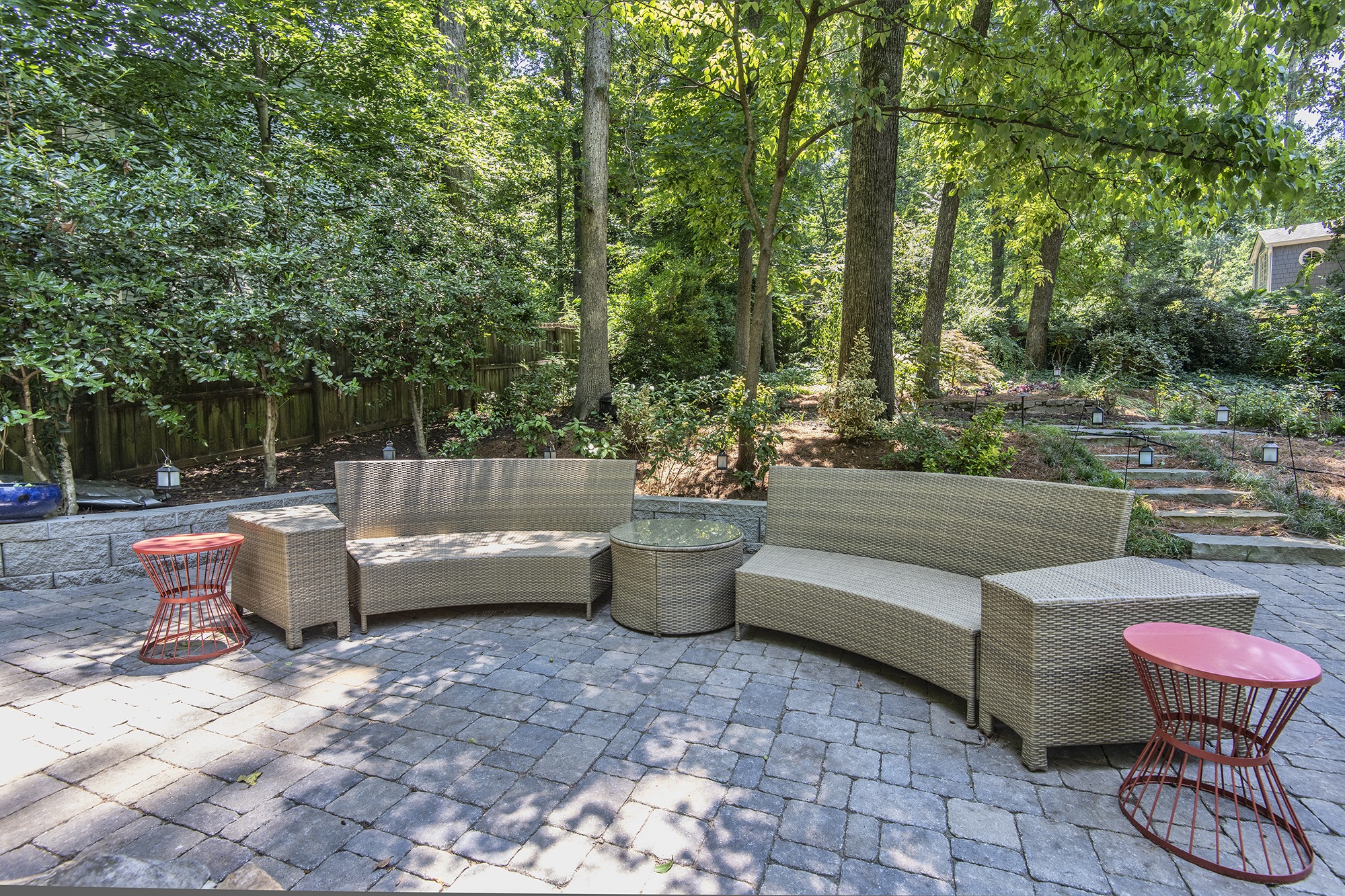 Outdoor patio features curved wicker seating, glass center table, and red side tables, surrounded by lush trees, greenery, and stone steps.