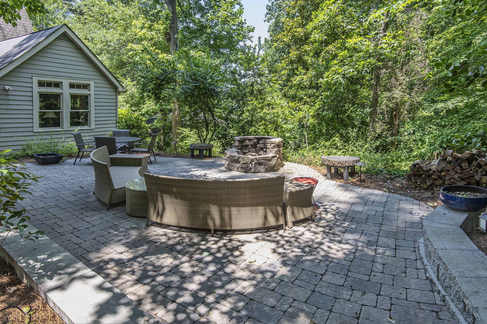 Stone patio with wicker seating, fire pit, and outdoor dining set, surrounded by lush greenery and a wooden house exterior in a serene setting.