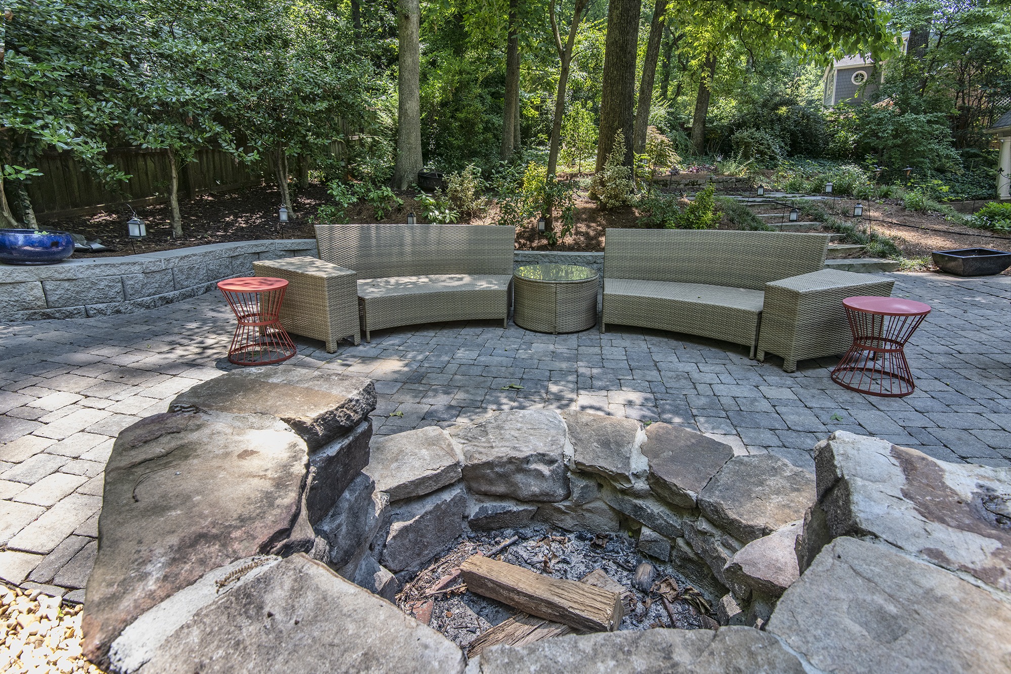 A cozy outdoor patio with stone fire pit, wicker seating, and lush trees creating a serene garden backdrop, perfect for gatherings.