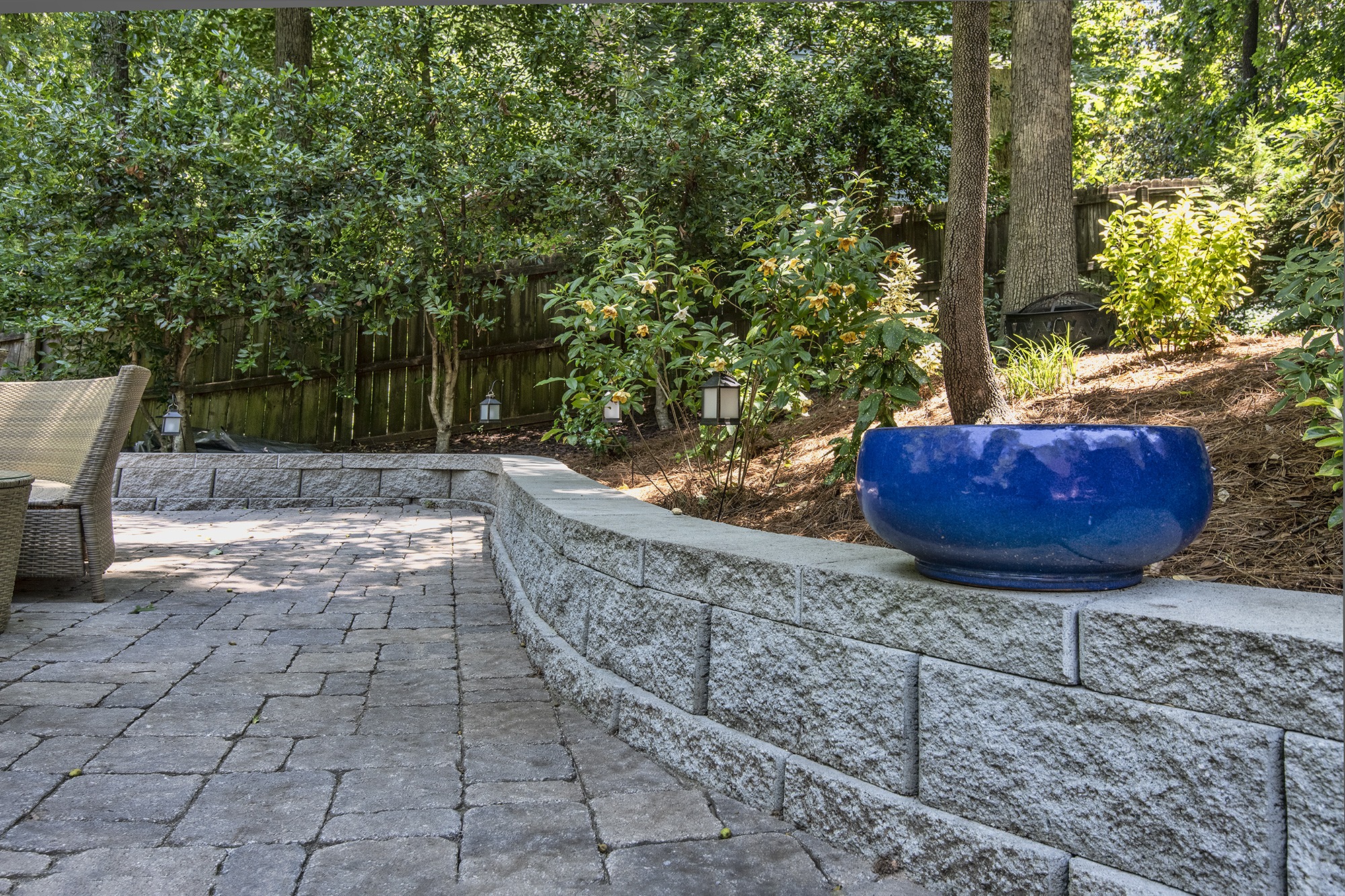 A serene garden with a curved stone wall, large blue planter, and lush plants, surrounded by patio seating and trees.