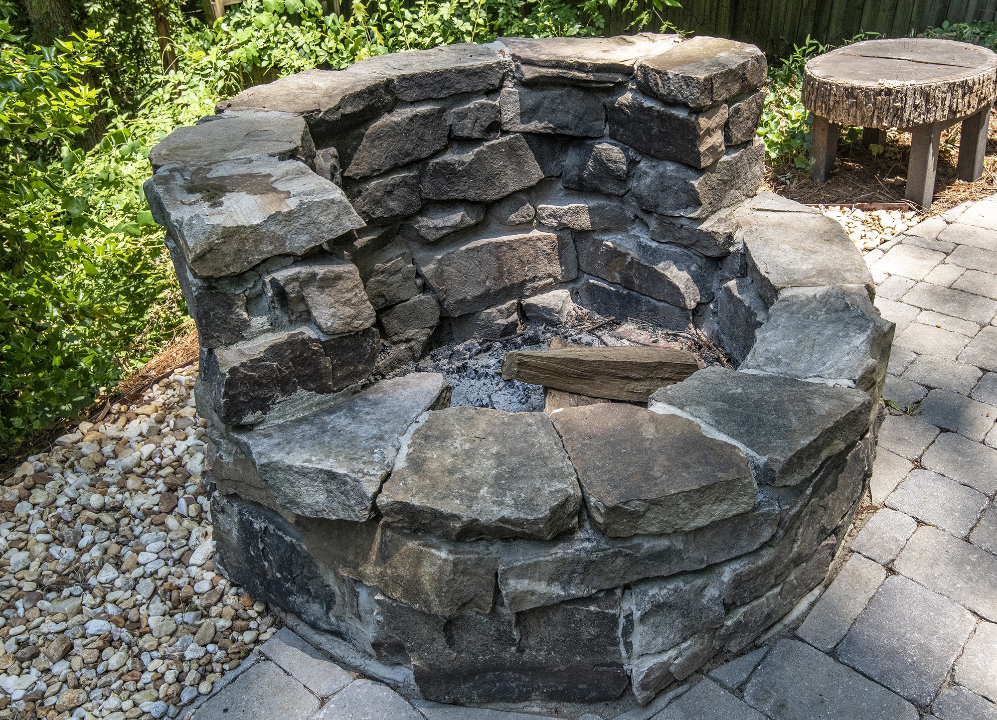 A rustic stone fire pit on a paved patio, surrounded by greenery and a tree stump table.