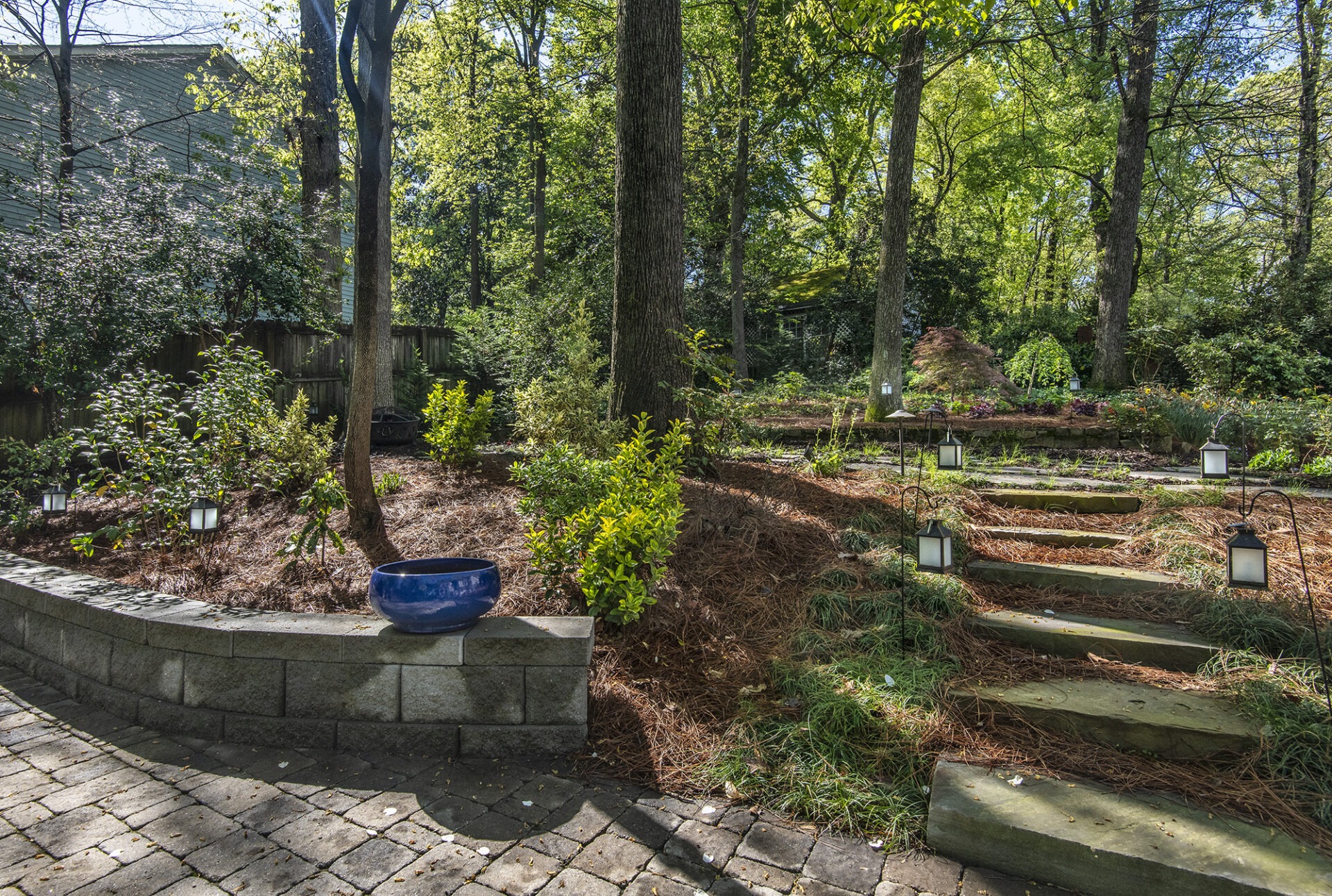 A serene garden features lantern-lit stone steps, lush greenery, and a blue pot on a brick wall, surrounded by tall trees.