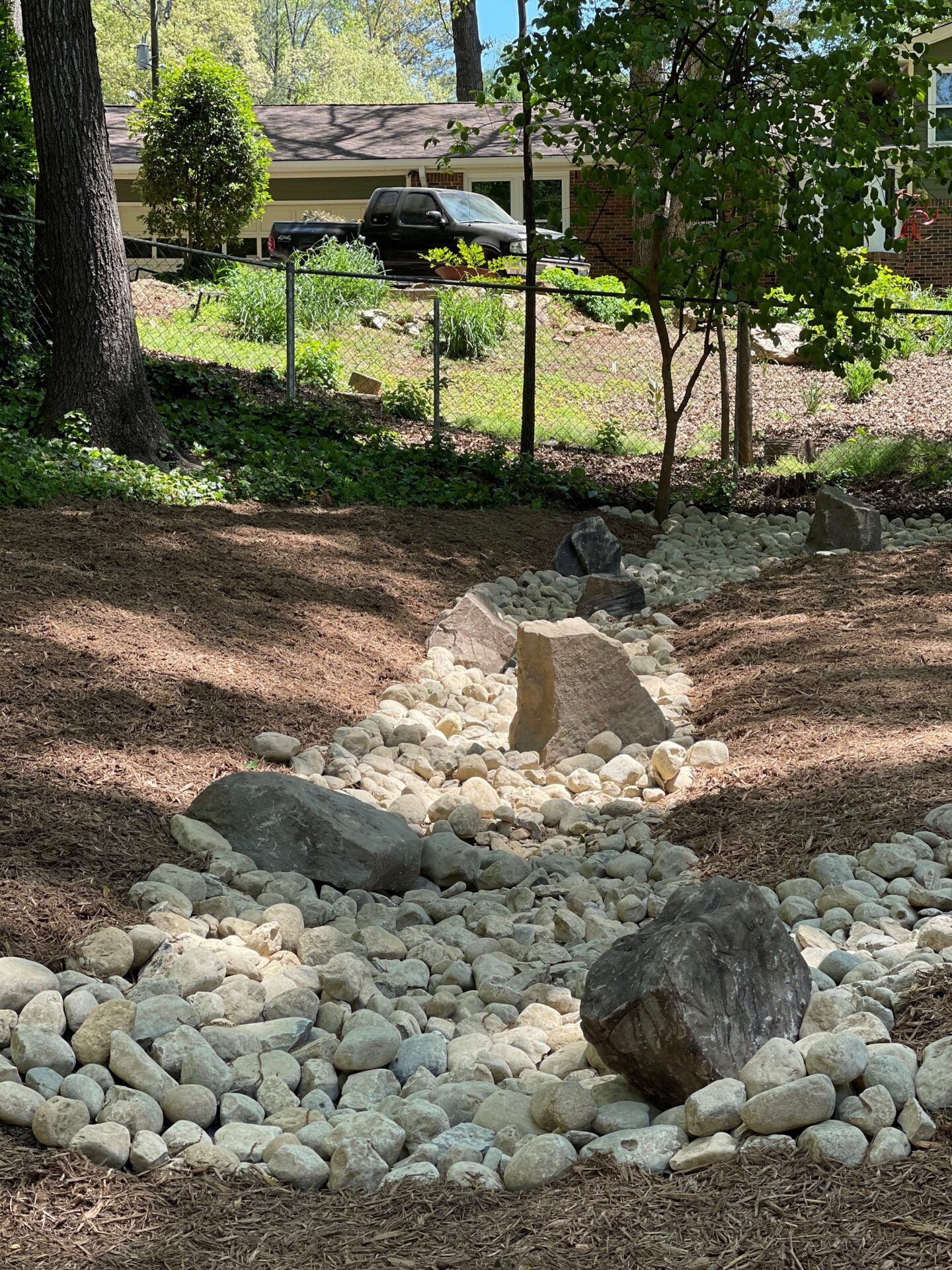 A dry creek bed with large rocks runs through a wooded backyard. A house and a parked vehicle are visible in the background.