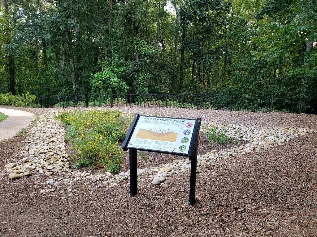 A rain garden with informative signage is surrounded by mulch, rocks, and greenery, set against a backdrop of dense forest trees.