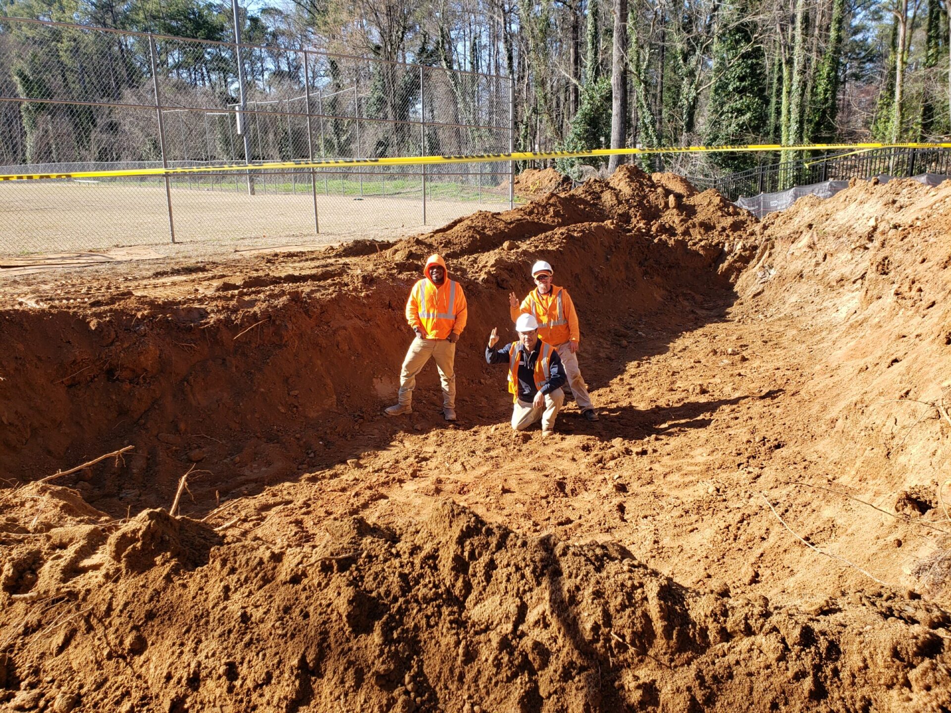 Three individuals in safety gear stand in a deep excavation site, bordered by a fence, surrounded by trees, under clear blue skies.