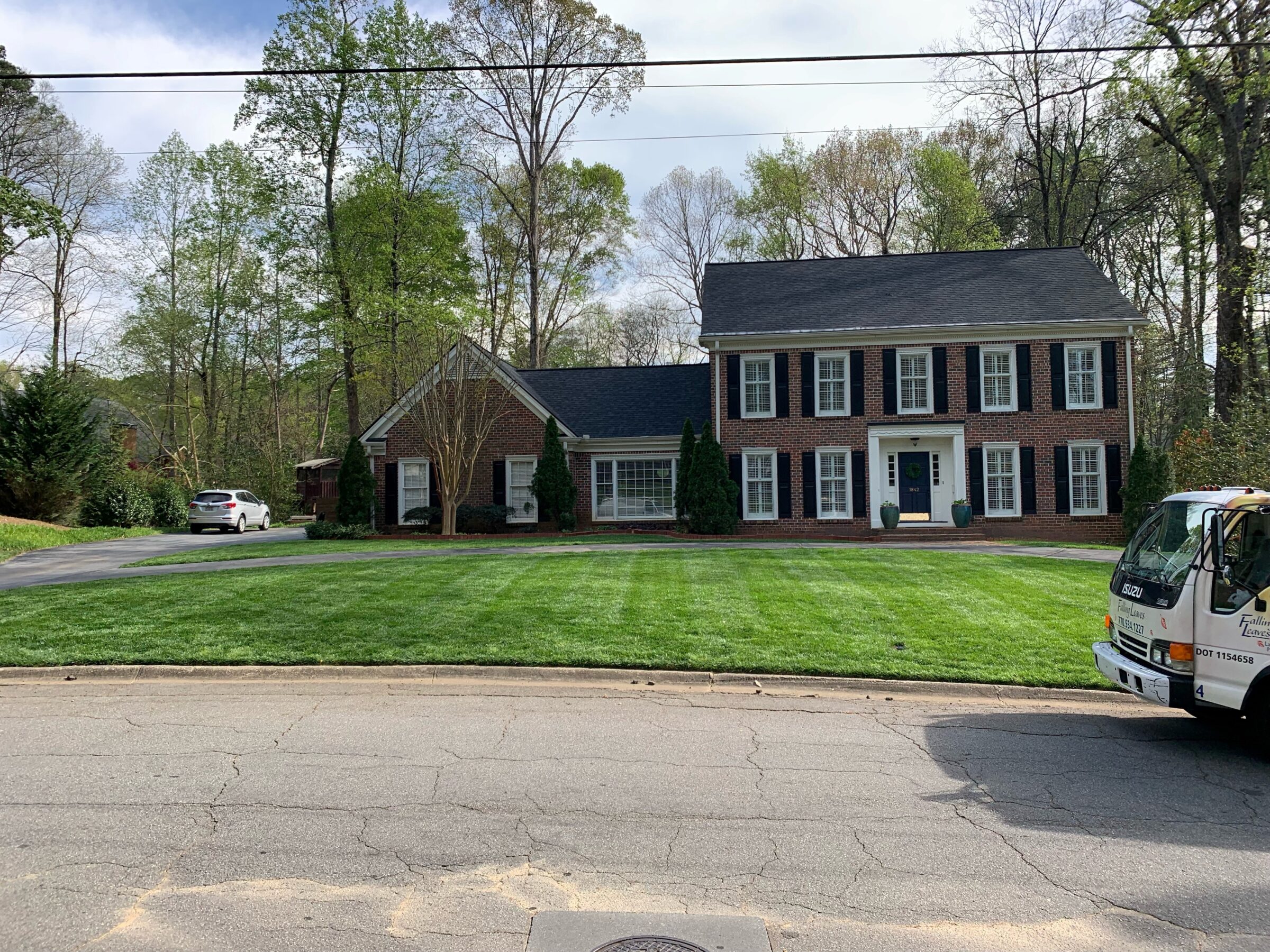A brick house with black shutters, surrounded by trees, cars parked outside, and a truck labeled "Isuzu" nearby on the street.